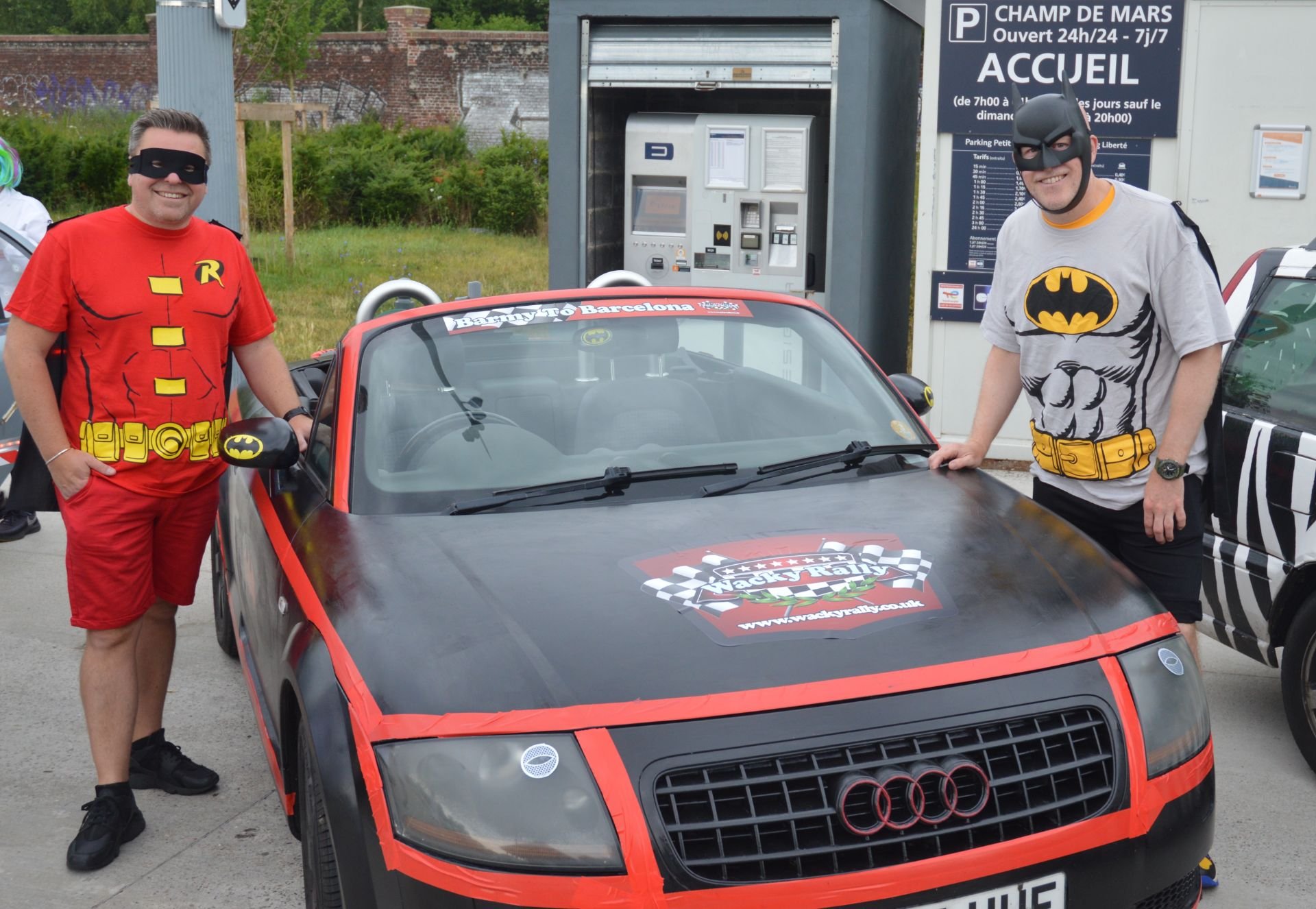 Two men dressed as comic book characters Batman and Robin standing next to a black and red race car, with a parking payment kiosk in the background.