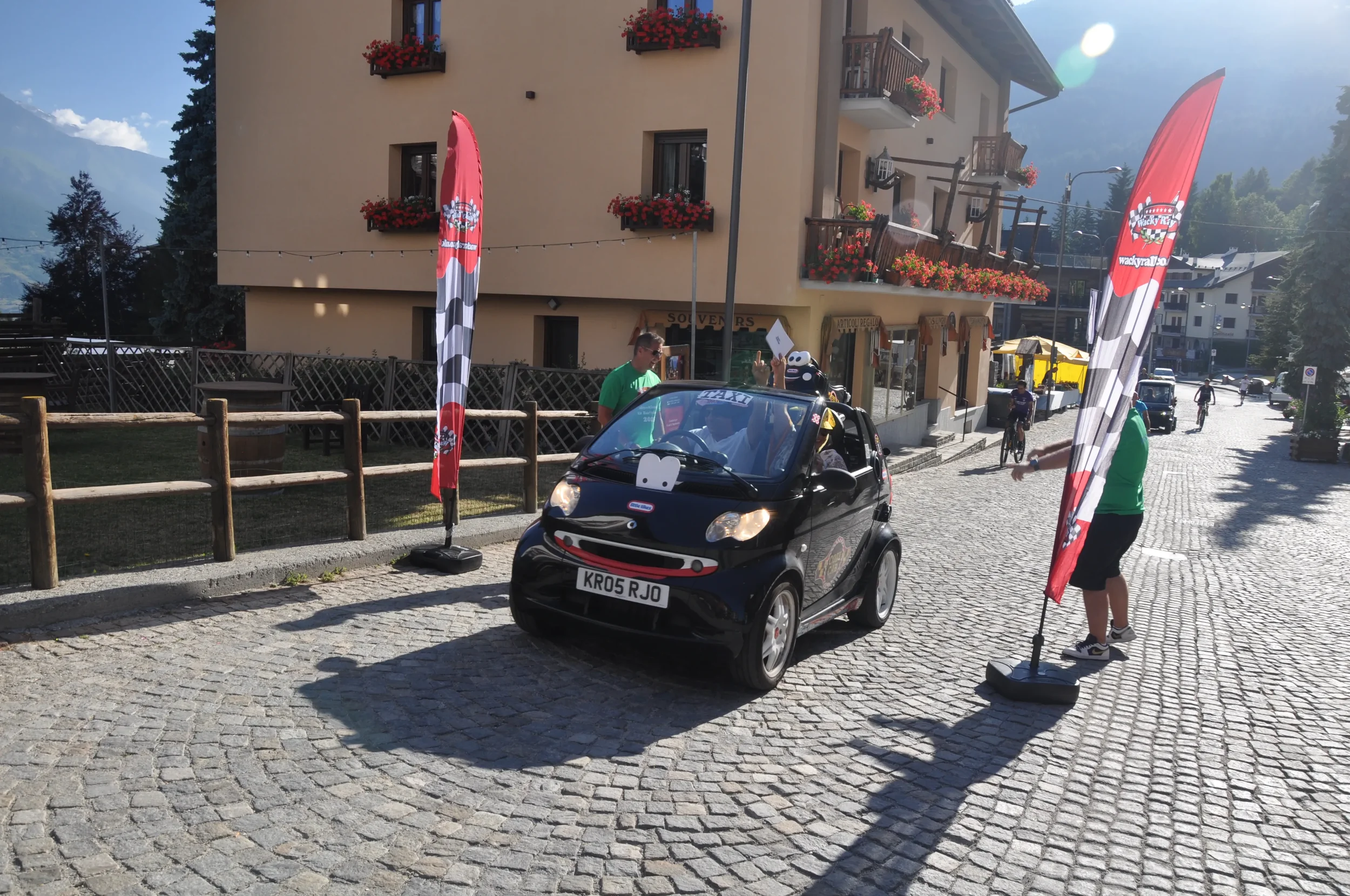 A small black Smart car decorated with a smiley face, with people around it waving and standing near two red and black flags with checkered patterns and a logo, in a cobblestone street in a mountain town with flowers and buildings.