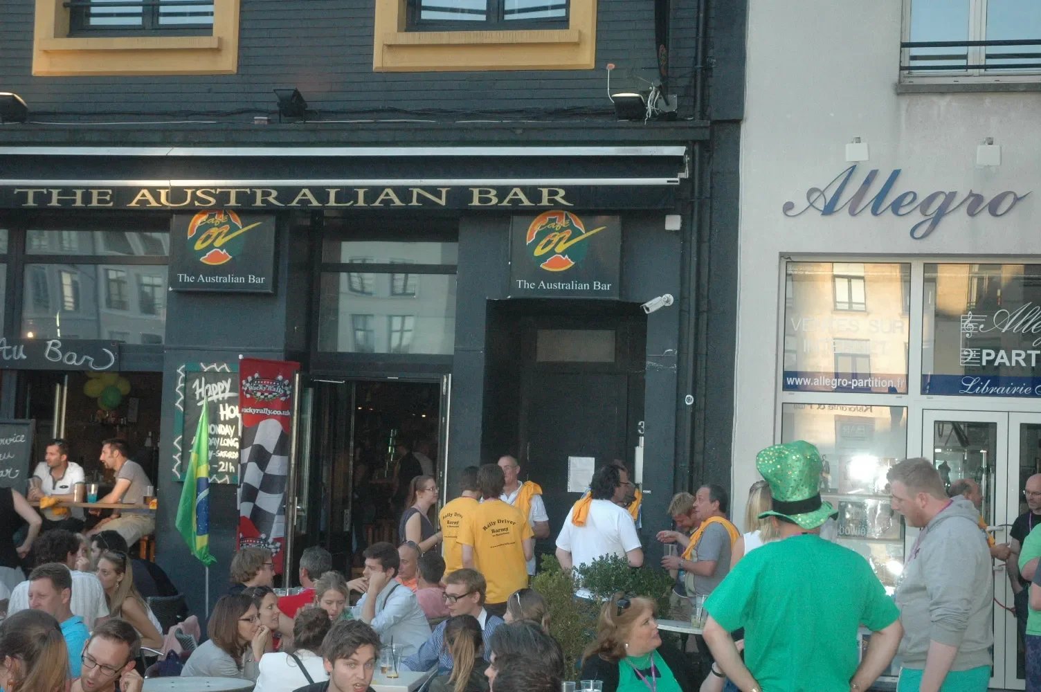 Crowd gathered outside The Australian Bar, a lively pub with people sitting and standing, wearing colorful clothing including green hats, in an urban setting with neighboring businesses.
