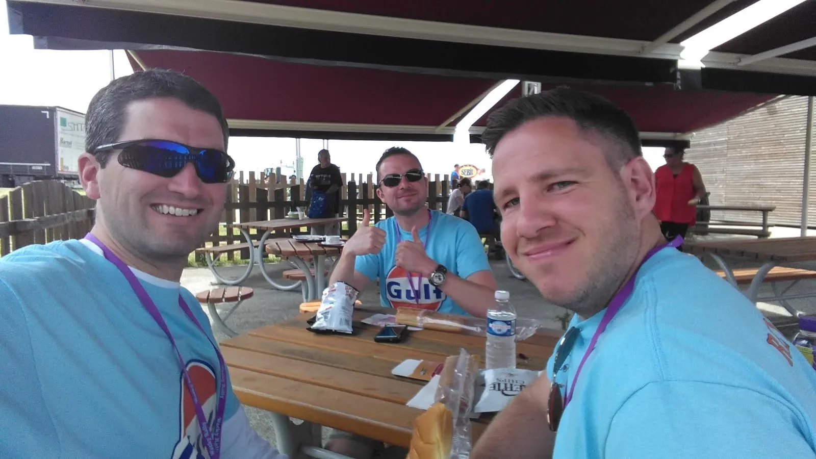 Three men sitting at a picnic table wearing blue T-shirts, with food and drinks on the table, smiling and posing for the camera