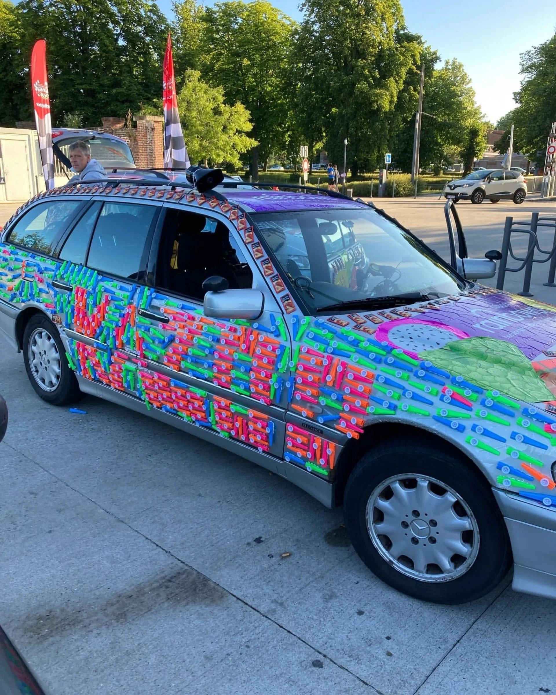 A silver station wagon decorated with colorful plastic straws and paper cutouts, with a pink and green heart-shaped design on the hood, and small flags on the roof. There is a person standing near the vehicle in an outdoor area with trees and other c