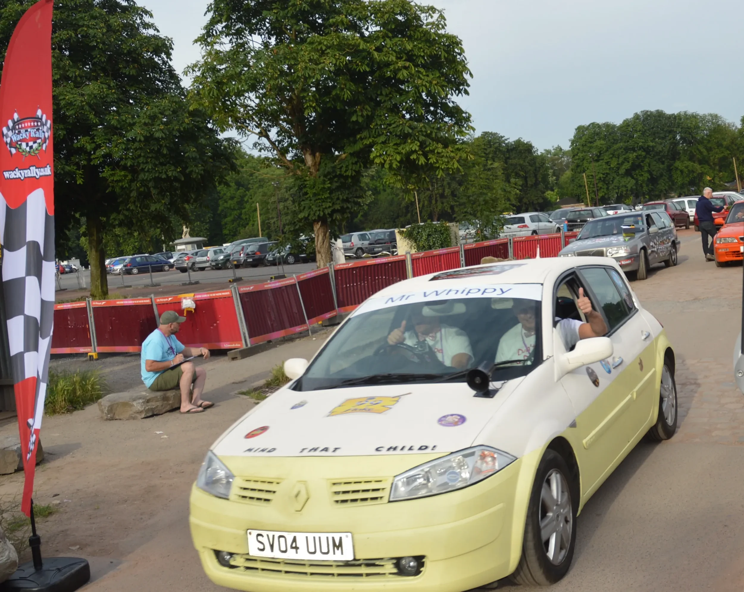 A yellow and white racing car with the words "Mr. Whippy" on the windshield and various stickers and phrases on the body, parked outdoors at a racing event with people and other cars in the background.