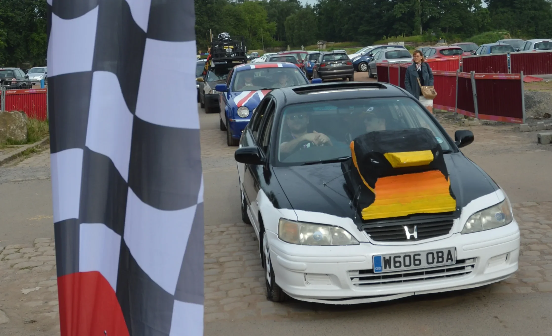 A black and white Honda car with a large, colorful, racing-style hood decoration on top, parked on cobblestone pavement at a racing event. Several other race cars and spectators are in the background, and a checkered flag is visible in the foreground
