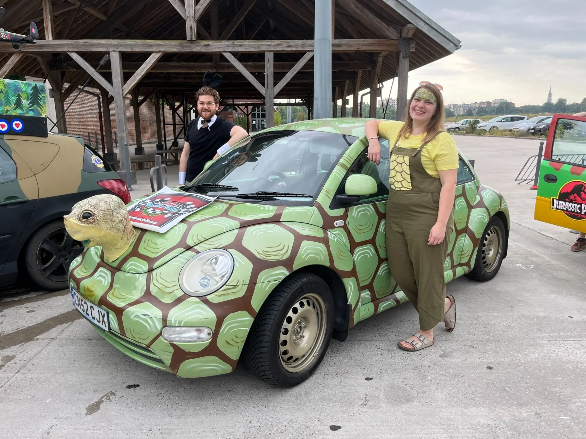 A woman dressed as a character from Jurassic Park and a man dressed as a bunny pose next to a small, eco-friendly, turtle-themed car with a turtle head attached to the front. They are in a parking lot with a wooden structure and other cars, some with