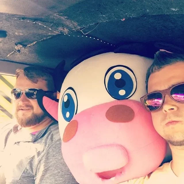 Two men taking a selfie with a large cartoon cow mascot. One man wears sunglasses and a gray jacket, the other wears sunglasses with pink reflective lenses and a yellow shirt. The cow mascot has big blue eyes, pink cheeks, and a pink snout.