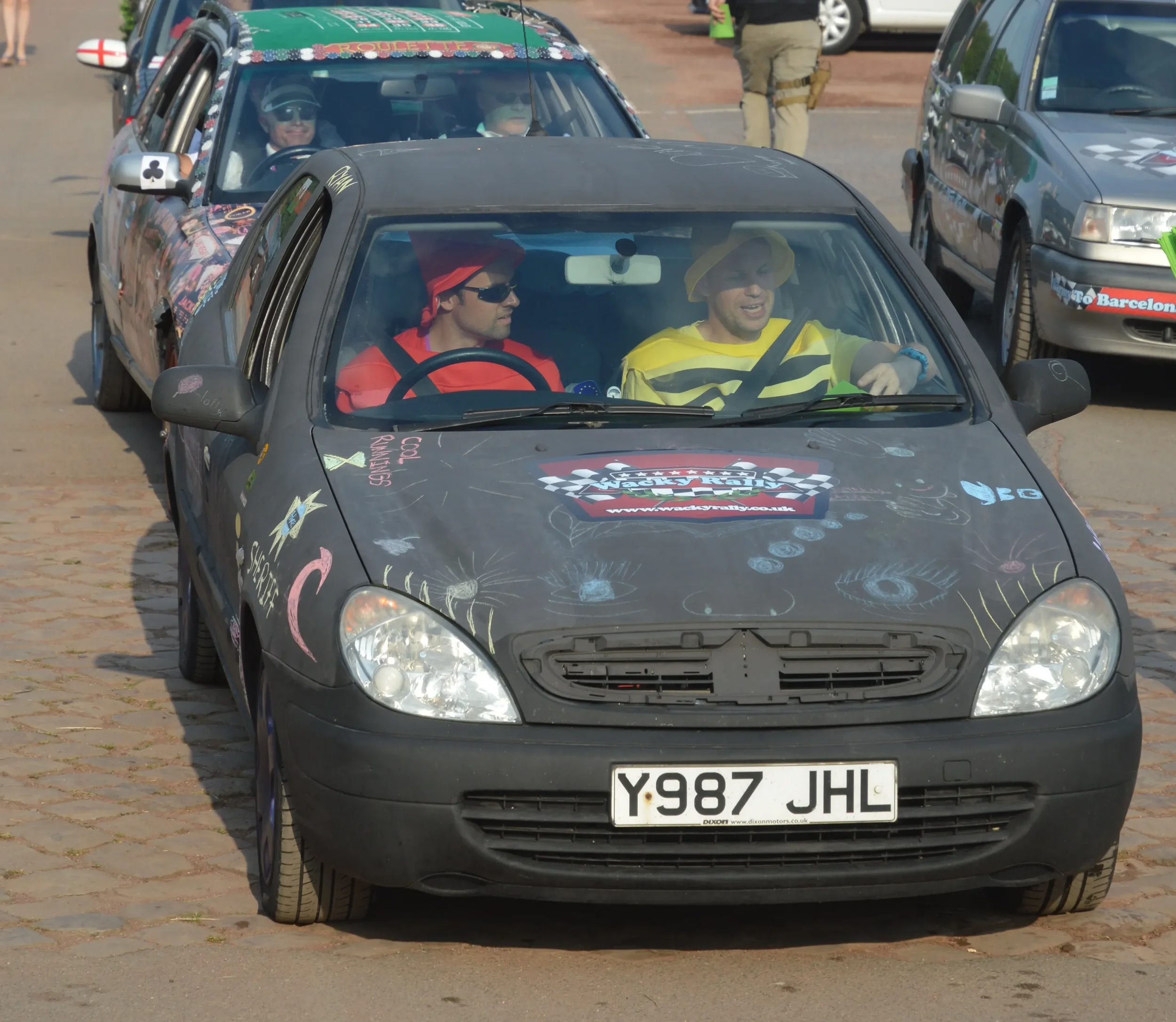 A black car with colorful chalk drawings on the hood and sides, carrying two men dressed as comedians with one in a yellow hat and striped shirt and the other in a red hat and sunglasses, parked on cobblestone pavement during a rally event with other