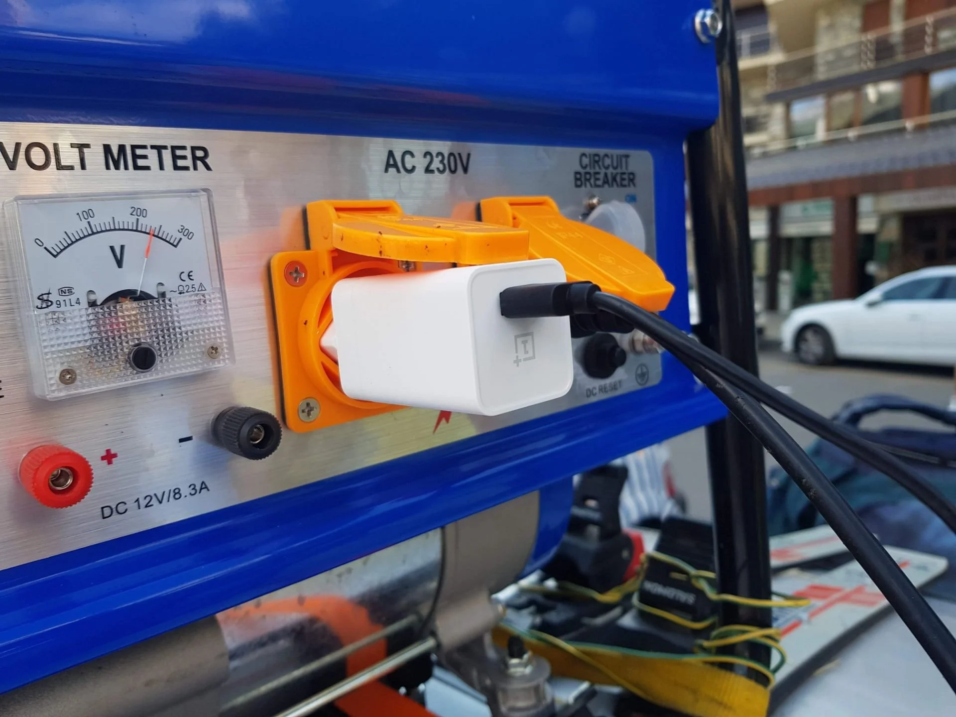 Close-up of a blue electrical device with a voltmeter, AC 230V label, circuit breaker, and a white charger plugged into an orange socket.