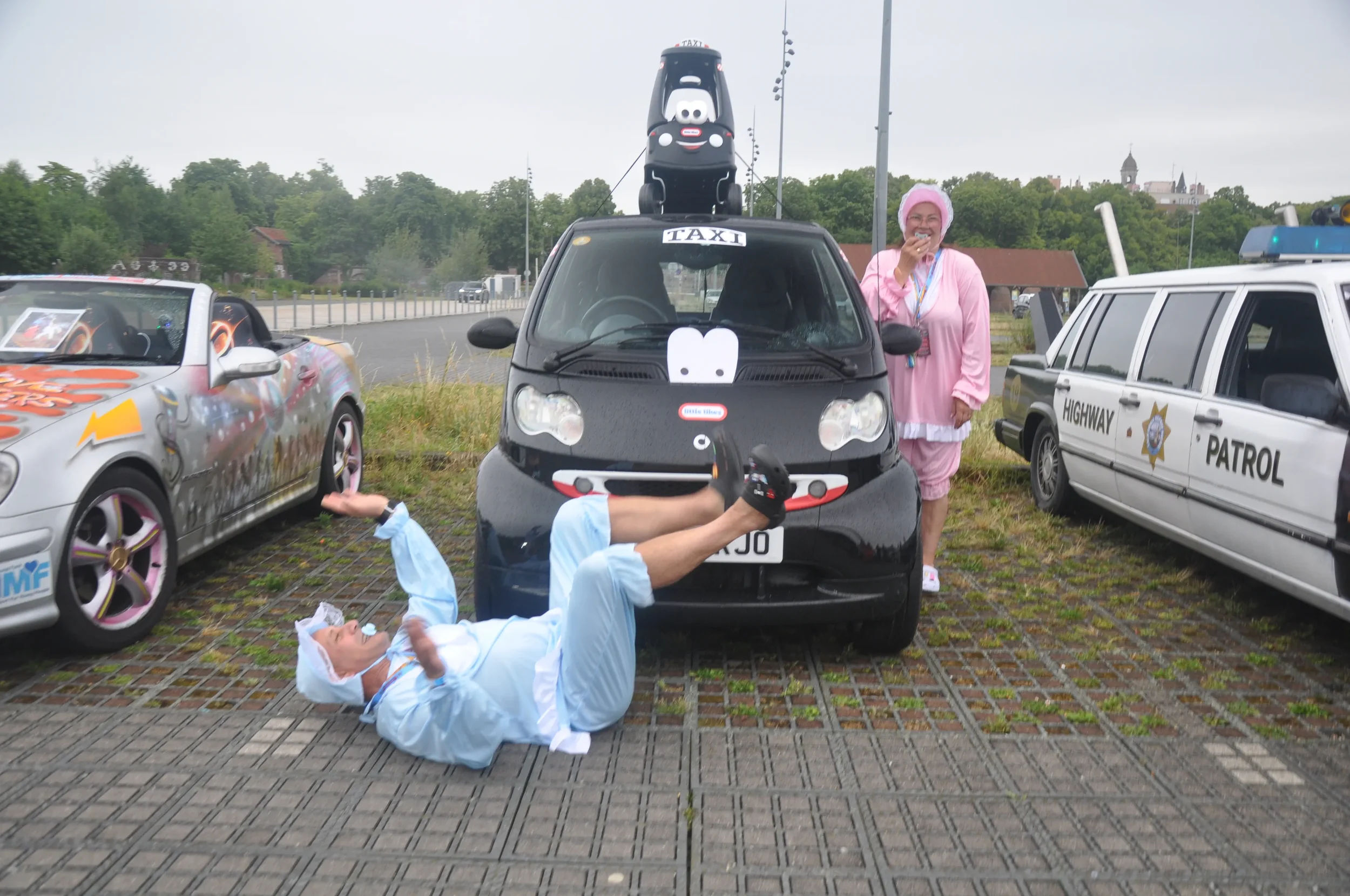 People dressed in costumes with cars decorated for a parade or event, including a person dressed as a healthcare worker lying on the ground with legs up, a woman dressed in pink with a nurse's outfit smiling, in front of a small black car with cartoo