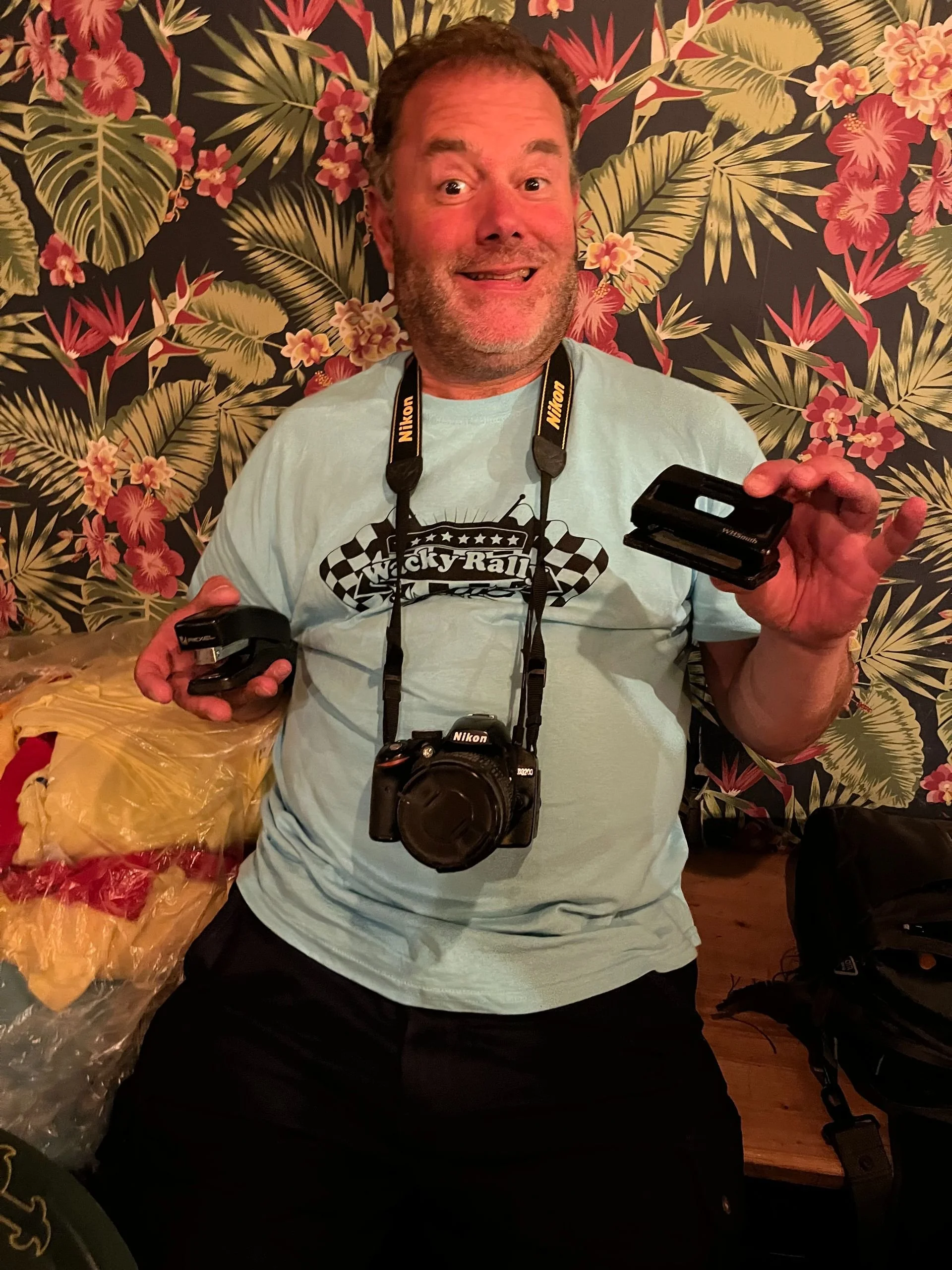 A man with short curly hair and a beard is sitting in front of a floral wallpaper, smiling and holding camera accessories. He is wearing a light blue T-shirt with a checkered race flag design and the words 'Lucky Rally' on it. Around his neck hangs a