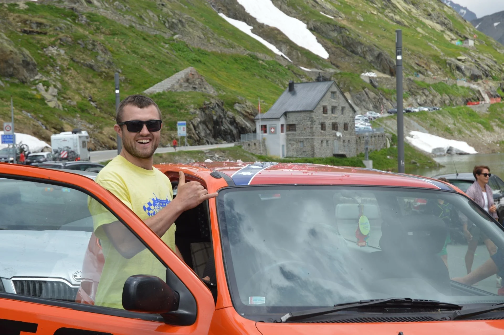 A smiling man wearing sunglasses and a yellow t-shirt, giving a thumbs-up while leaning out of the window of an orange car with a mountain and a building in the background.