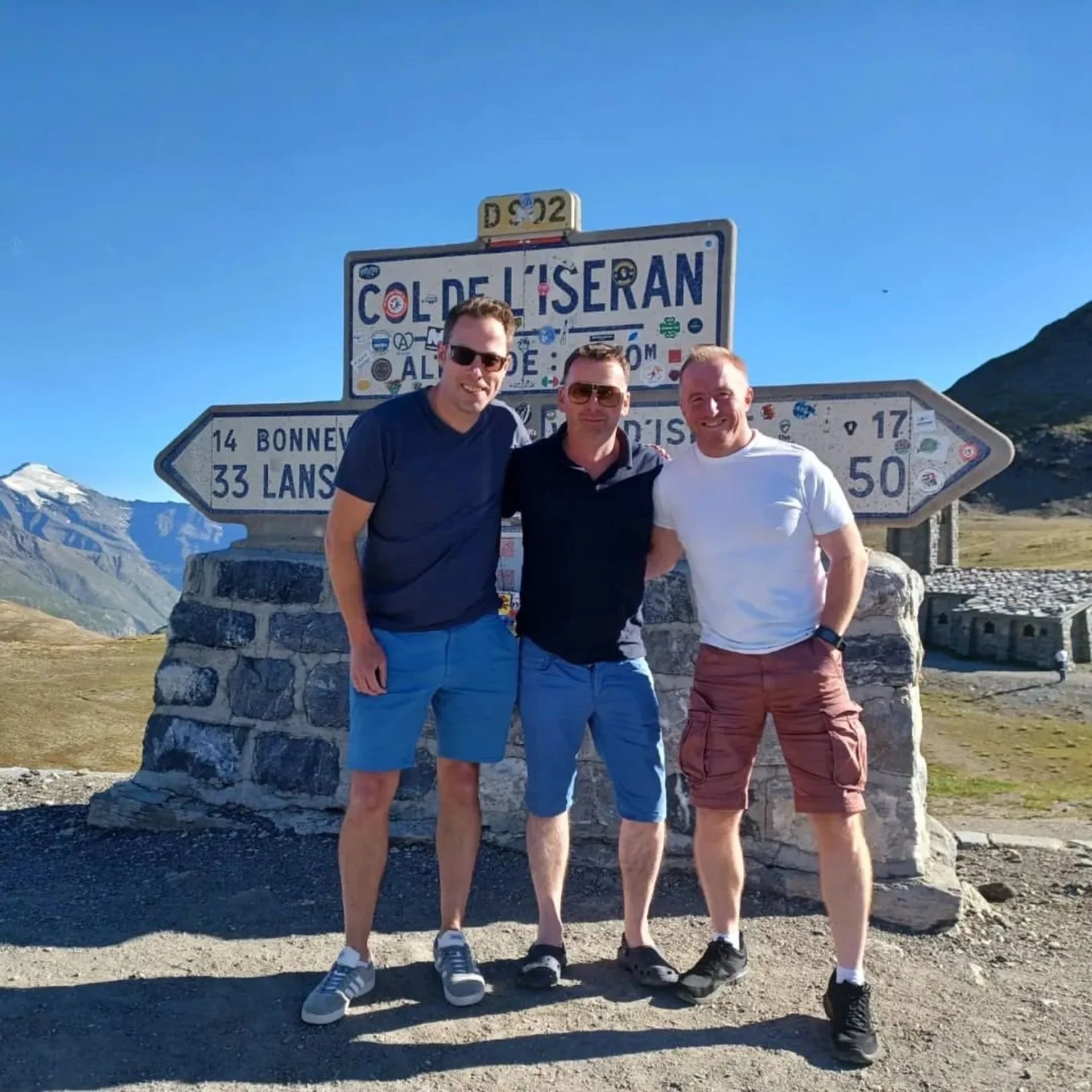 Three men standing together outdoors in front of a directional sign with mountains in the background, smiling at the camera.