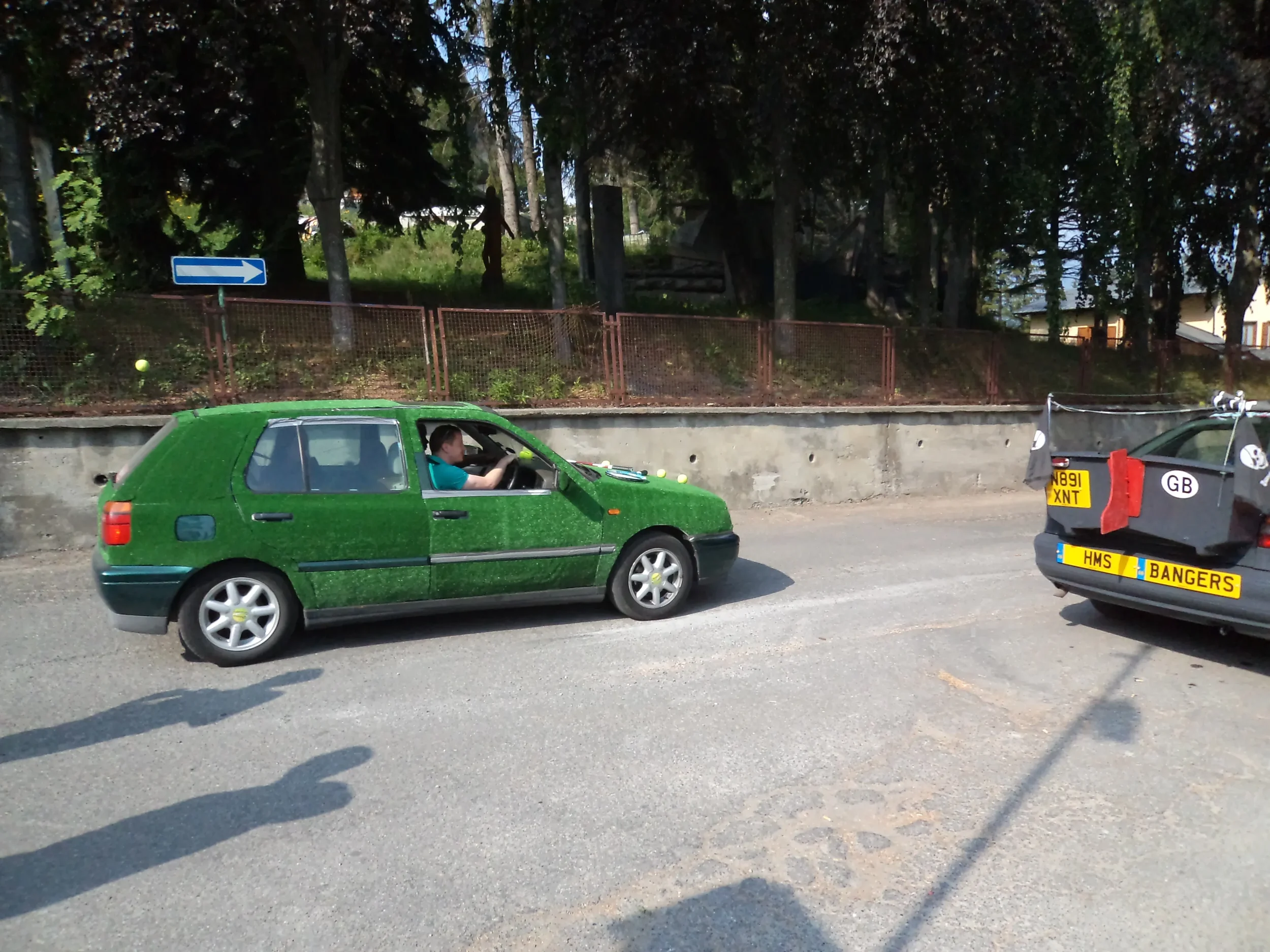 A green car with yellow tennis ball decals on the hood is parked on the side of the road. A person is inside the car, looking out the window. Next to it is a black car with a UK license plate, marked as a police vehicle with a yellow strip reading 'H
