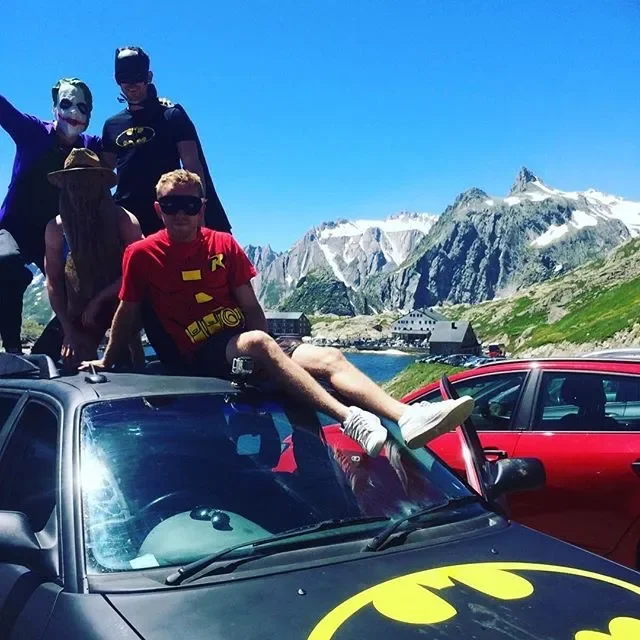 Group of people posing on top of a car with a Batman logo, with a mountain landscape and a lake in the background, on a sunny day.
