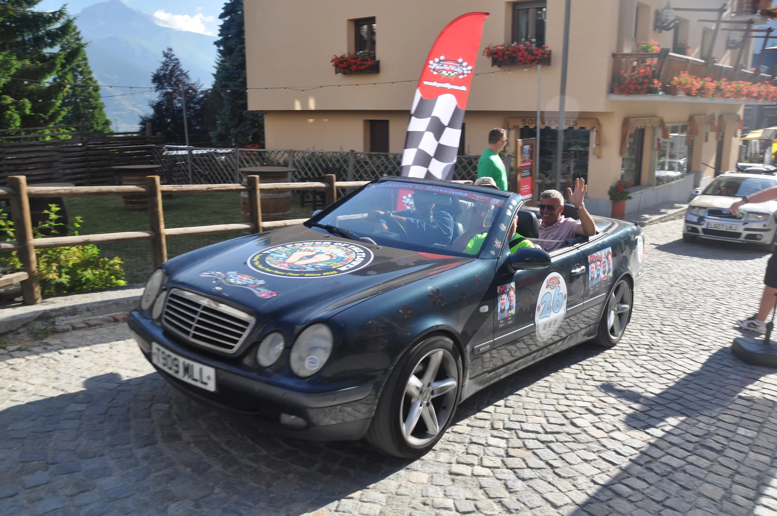 A black convertible car decorated with racing stickers and logos, carrying two people who are waving, driving on a cobblestone street in a European town.