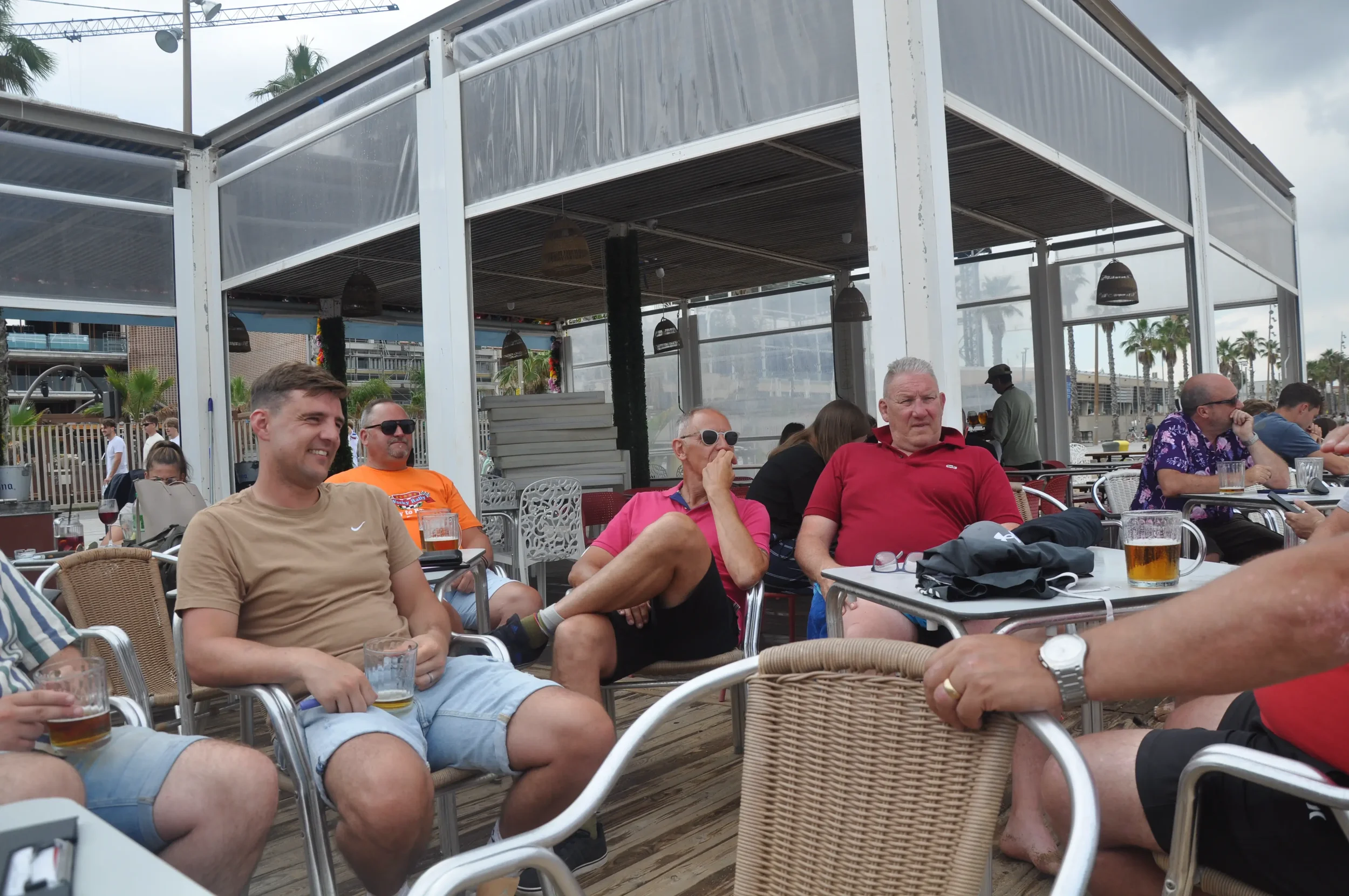 Group of people sitting at outdoor tables at a patio bar, enjoying drinks and casual conversation, with a modern, open-air structure in the background and palm trees in the distance.