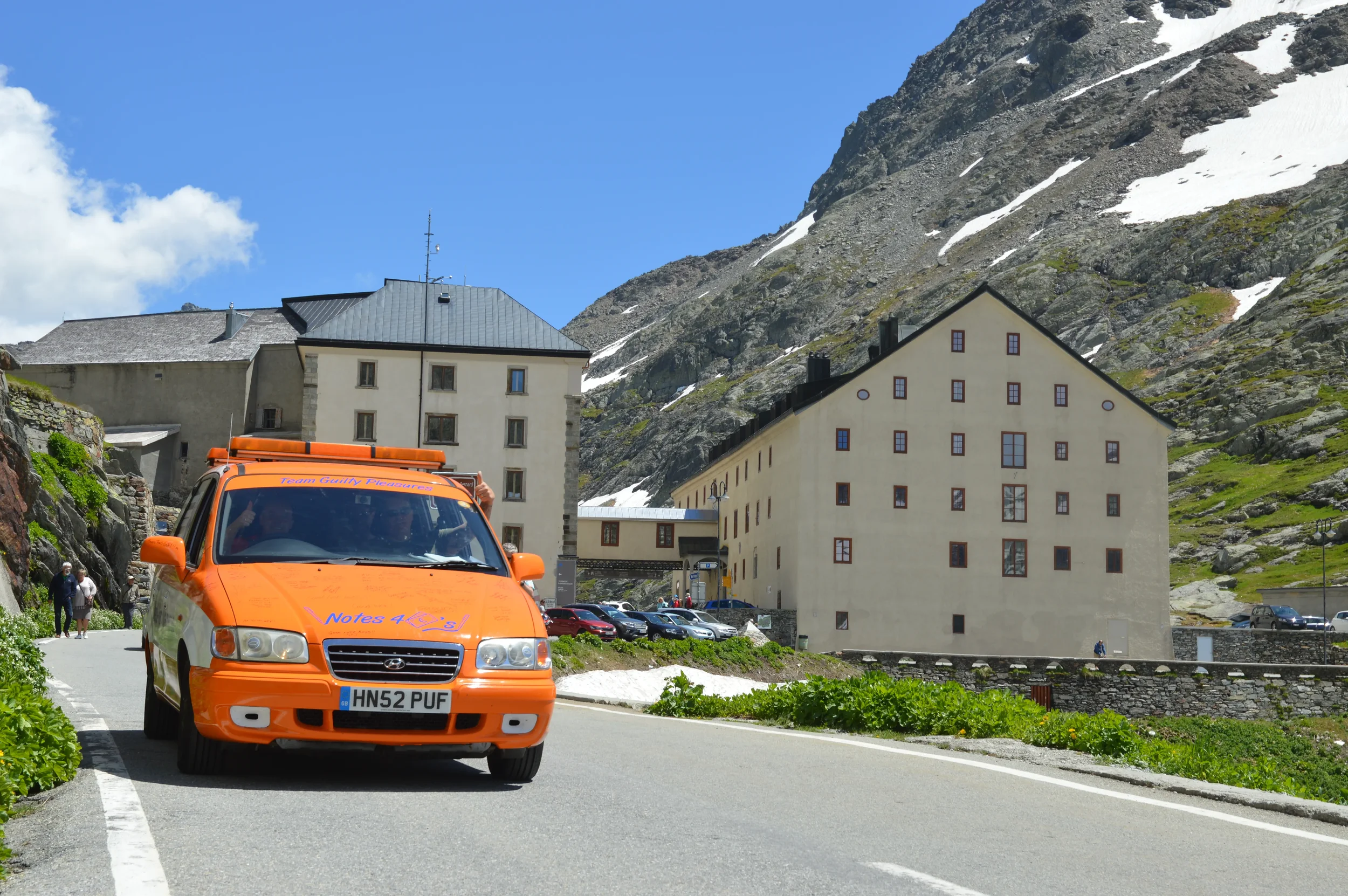 An orange Hyundai taxi with a license plate HN52 PUF driving on a mountain road in front of beige hotel buildings with snow patches on the mountainside.