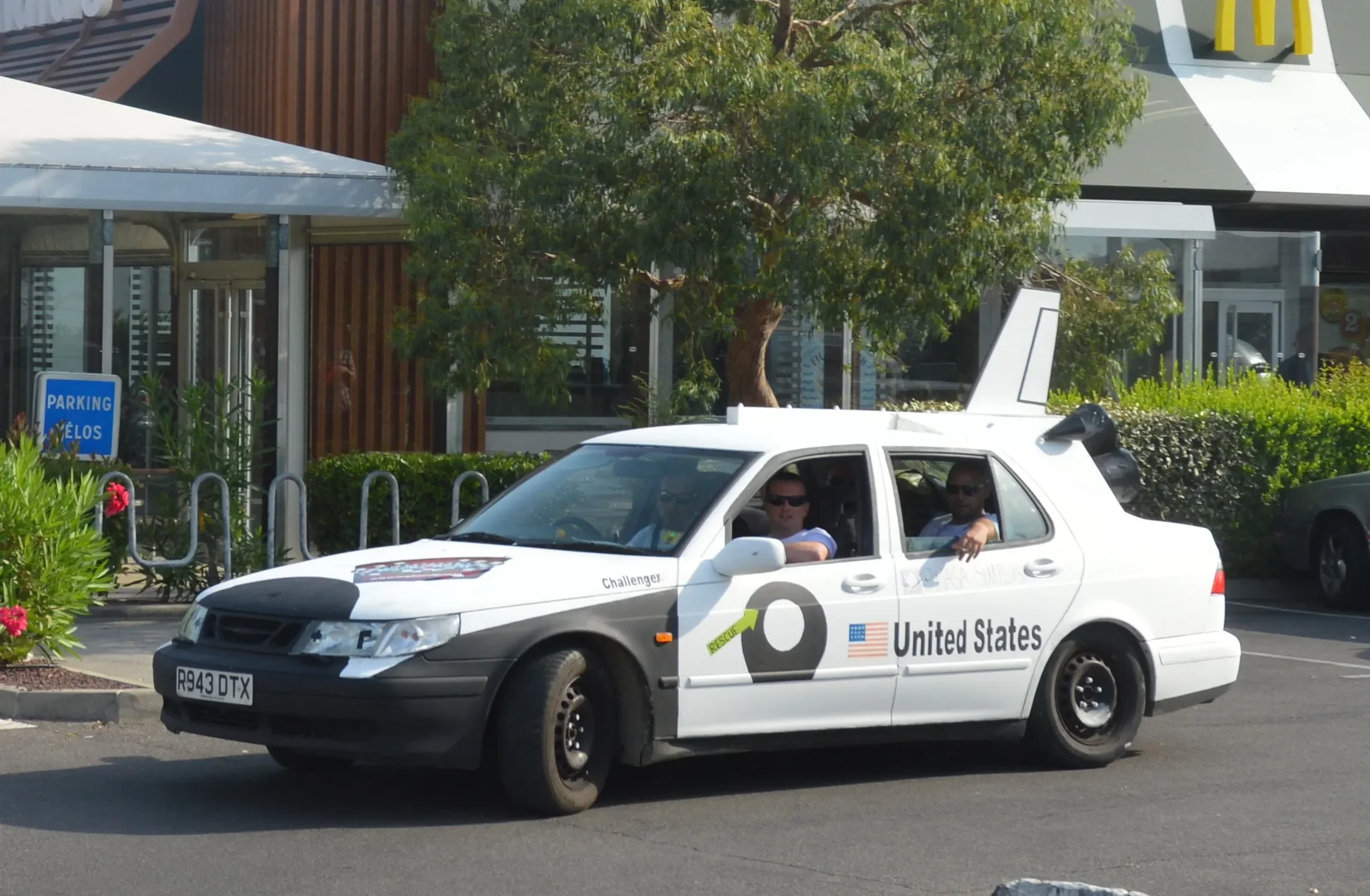 Amphibious vehicle designed to resemble a United States military rescue car, parked outside a commercial building with a tree and building in the background.