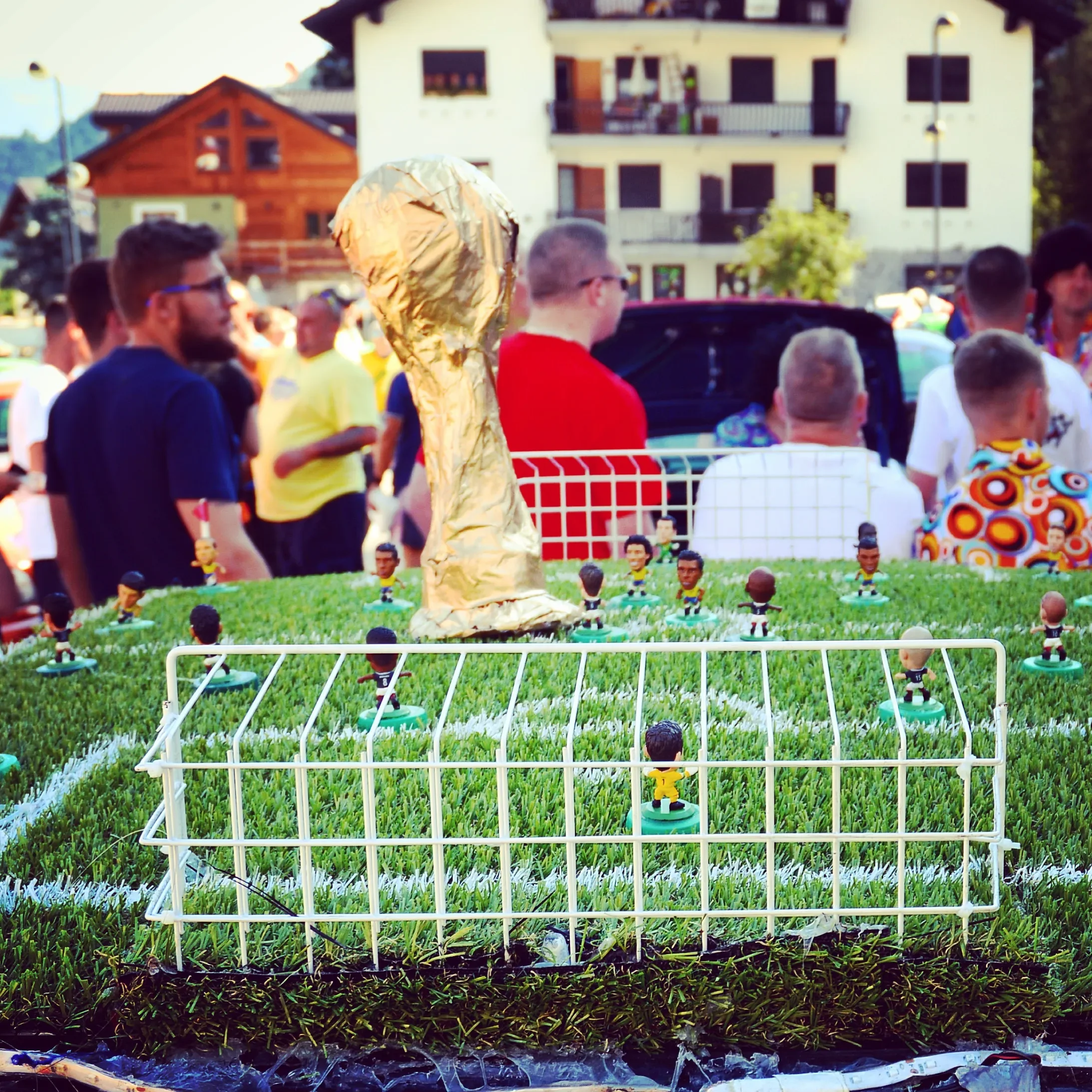 Miniature soccer field with small figurines and a large golden trophy on top, set on a grassy surface, with a crowd of people in the background.