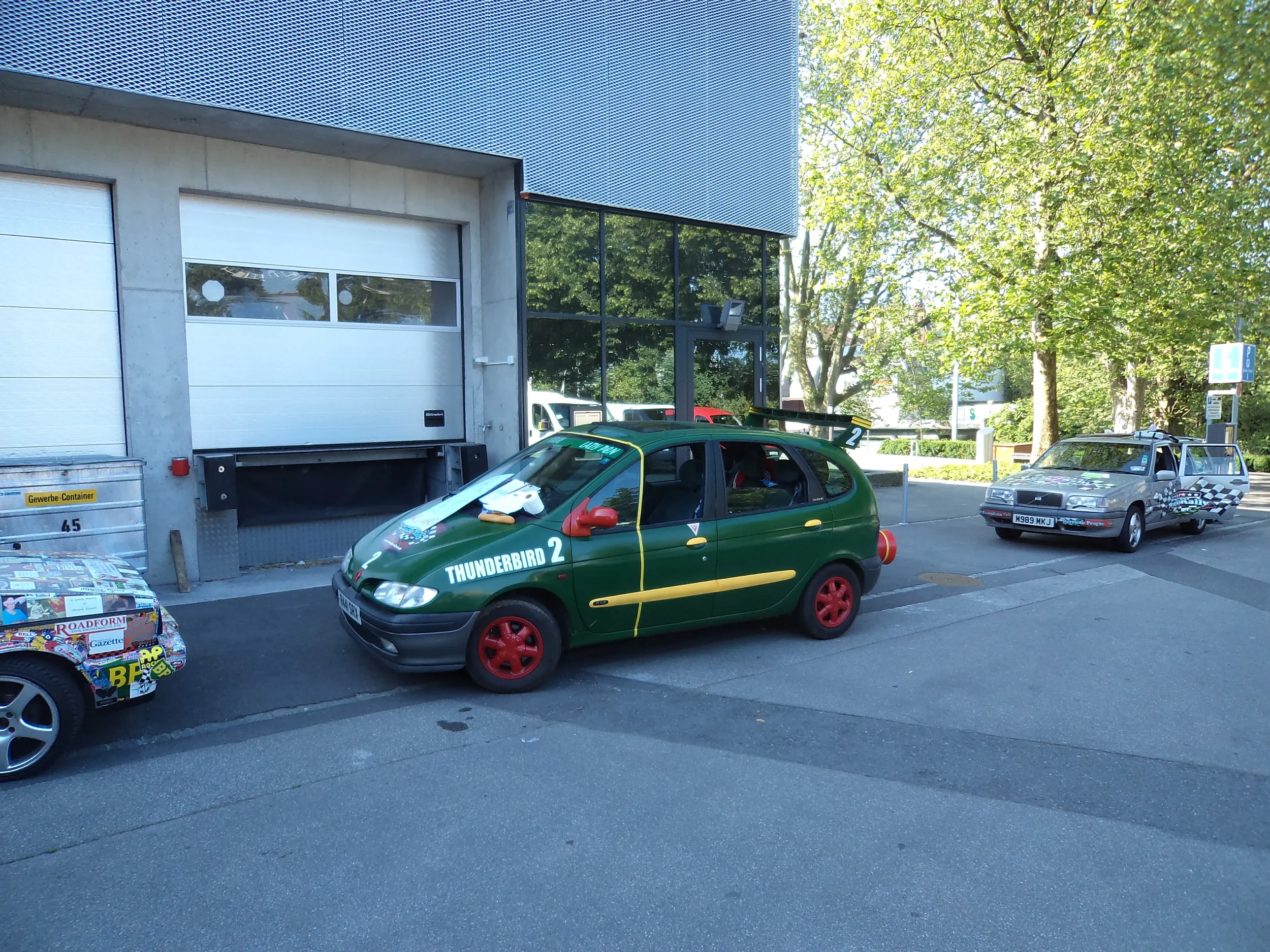 Three cars parked outside a building, with the middle car painted green and marked "Thunderbird 2." The car has red wheels and a large rear spoiler, and the front has a white paper and orange item on the windshield. The right car is silver with racin