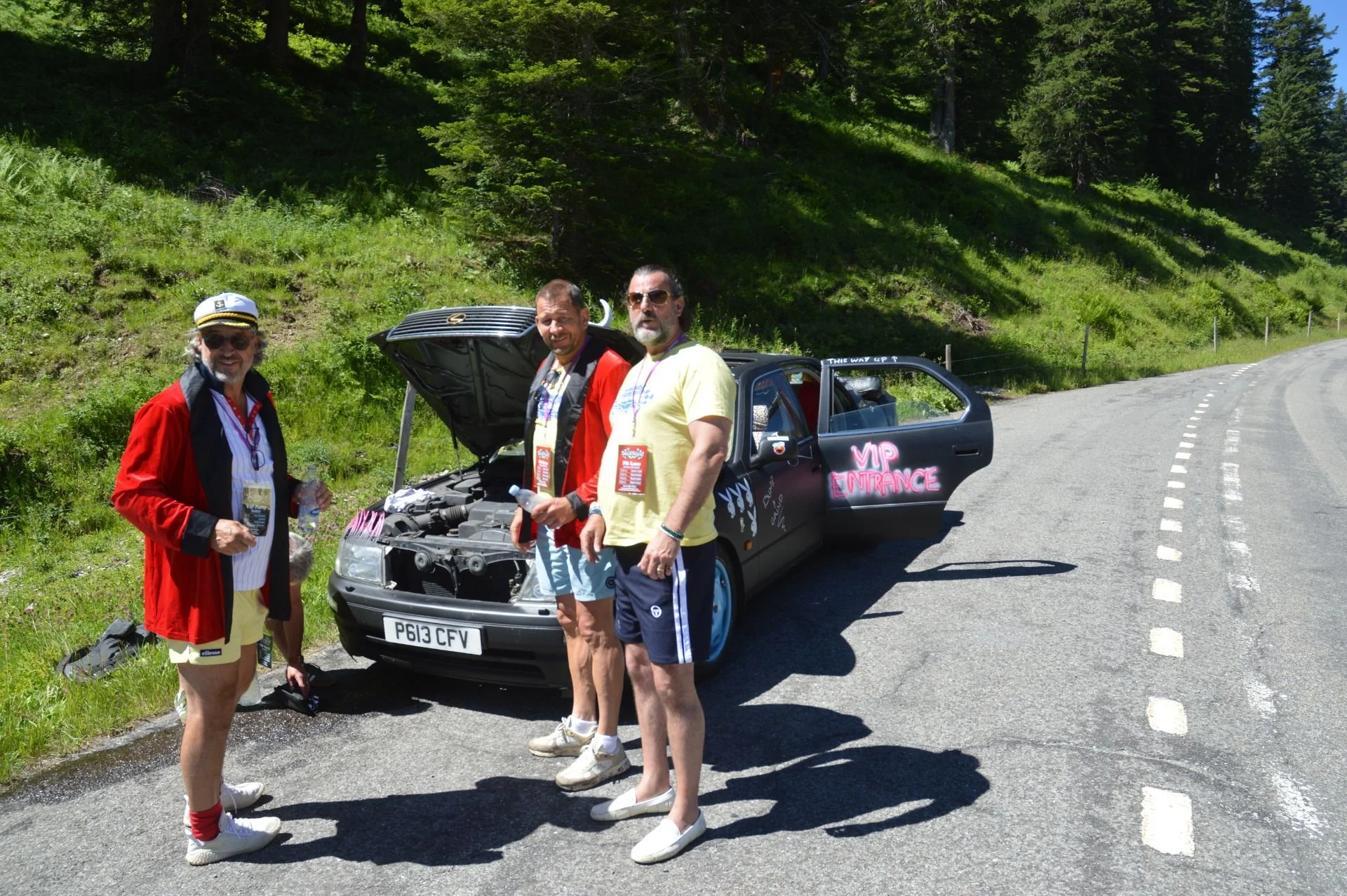 Three men standing next to a black car with its hood open, on the side of a winding mountain road. The car has pink signs reading 'VIP Entrance' on the door. The men are dressed casually, and the background features green trees and a grassy hillside.