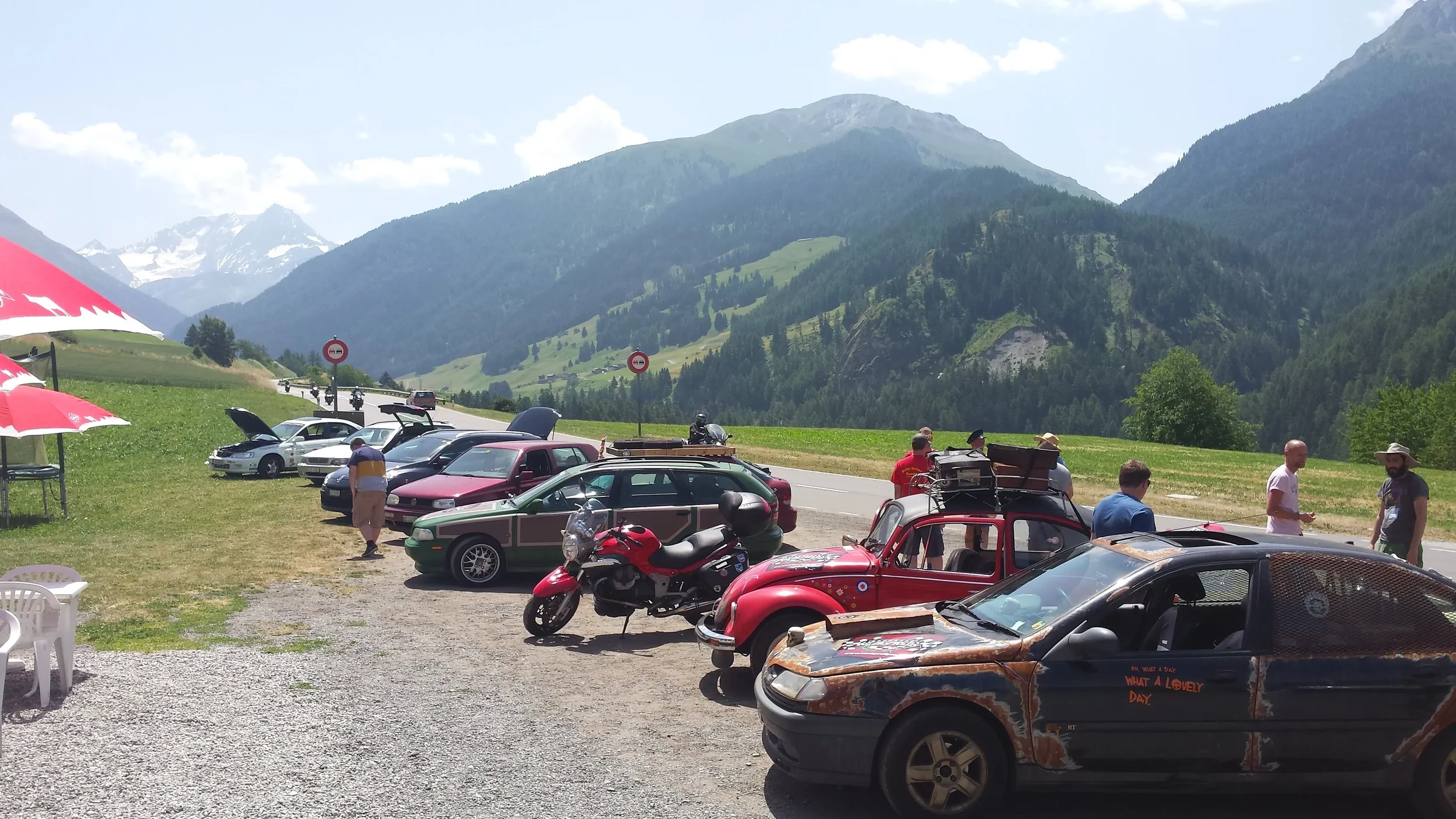 A scenic mountain view with a parking area filled with vintage and regular cars, a motorcycle, and people enjoying the sunny day among green hills and snow-capped mountains in the background.