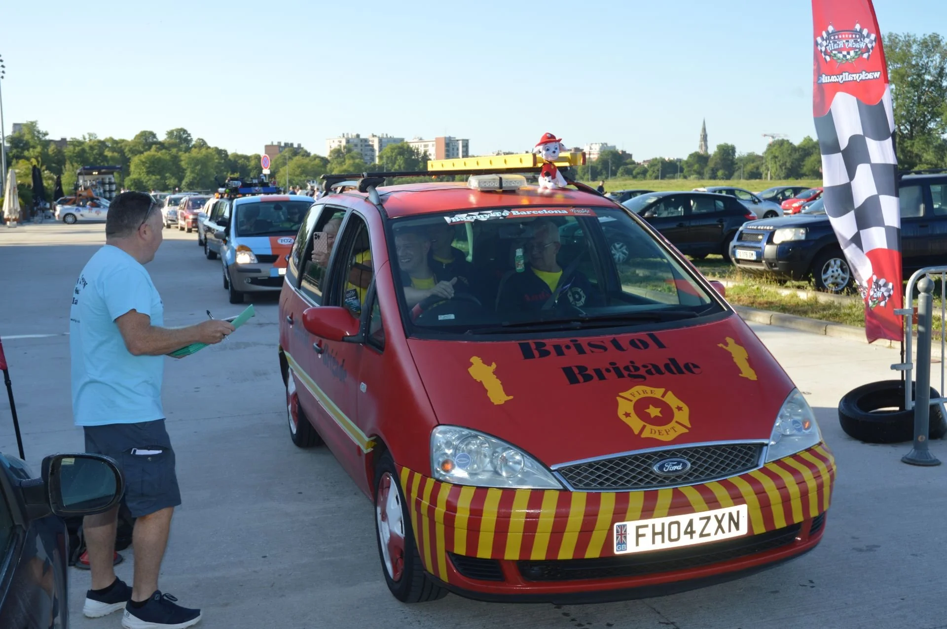 A red Ford car with 'Bristol Brigade' and fire department emblems on the hood, decorated with yellow and red stripes, parked outdoors in a lot with other vehicles. There is a man standing nearby holding a notebook, and two people inside the car smili