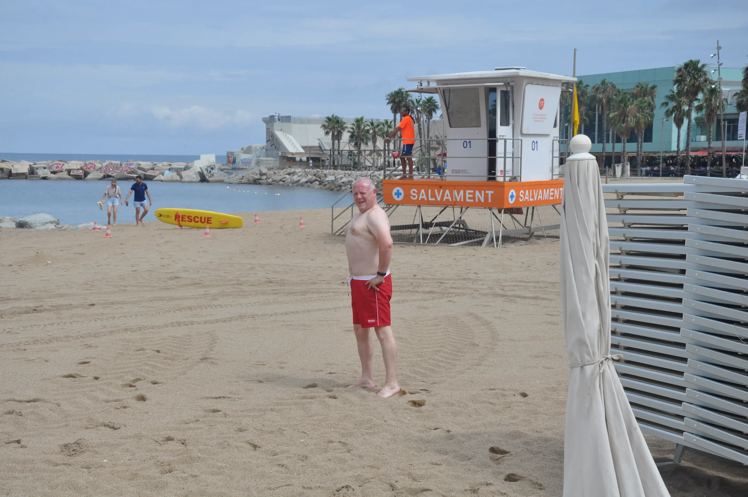 A man with white hair and red shorts standing on a sandy beach near a lifeguard tower, with two people walking near the water, and a yellow rescue board on the beach, under a partly cloudy sky.