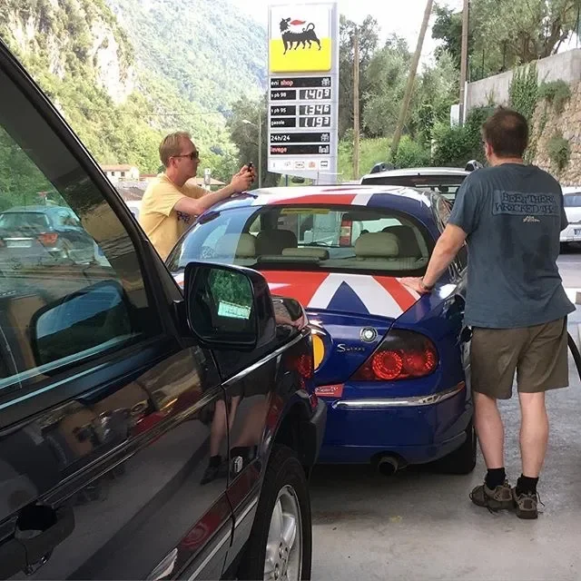 Two young men at a gas station, one taking a photo of the other who is standing next to a blue car with a Union Jack design on the back, with a petrol price sign in the background.