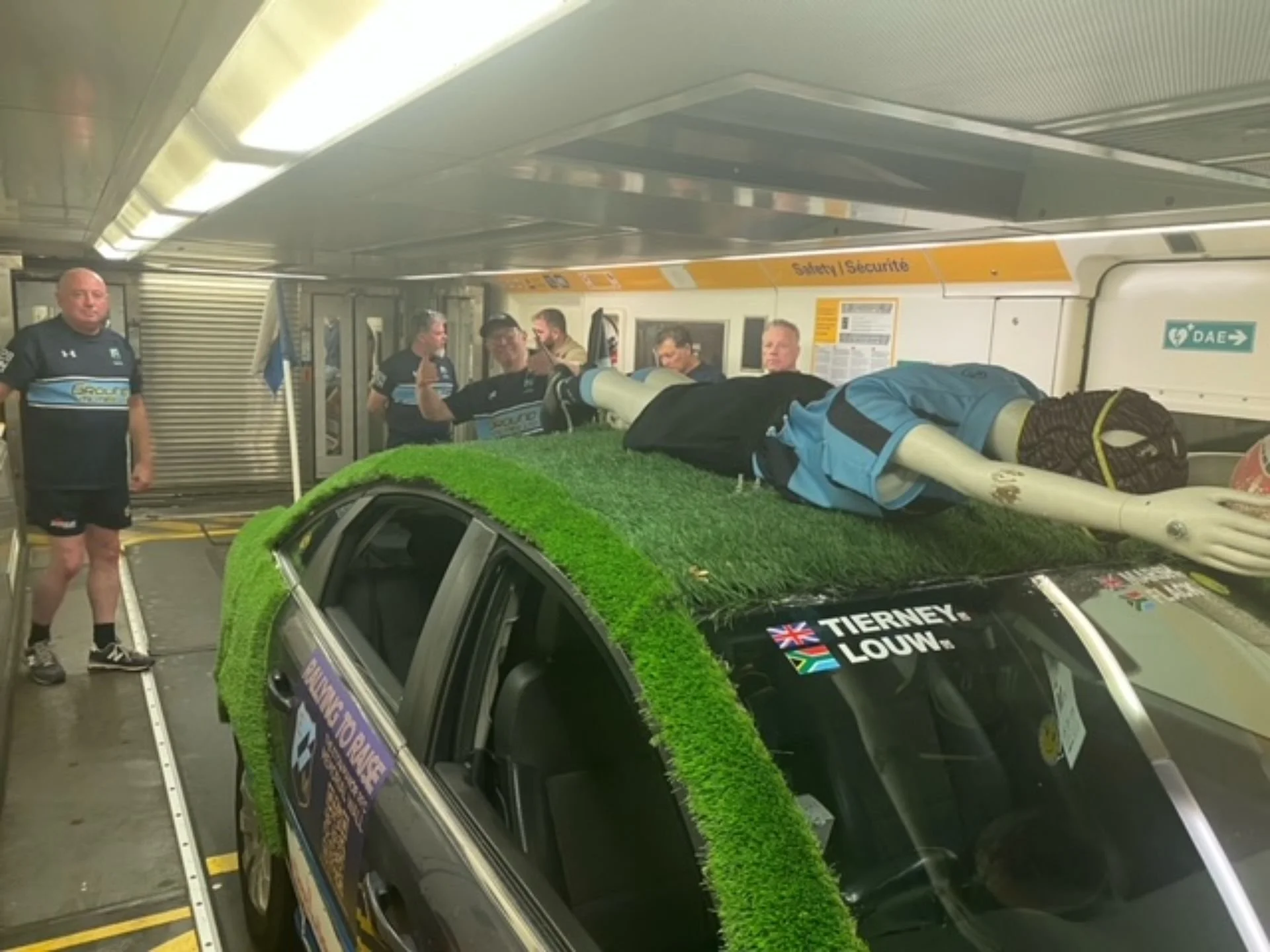 A car covered with artificial grass on the roof, with a mannequin in sports clothing lying on top, and several people in sports uniforms standing around inside an airport tunnel