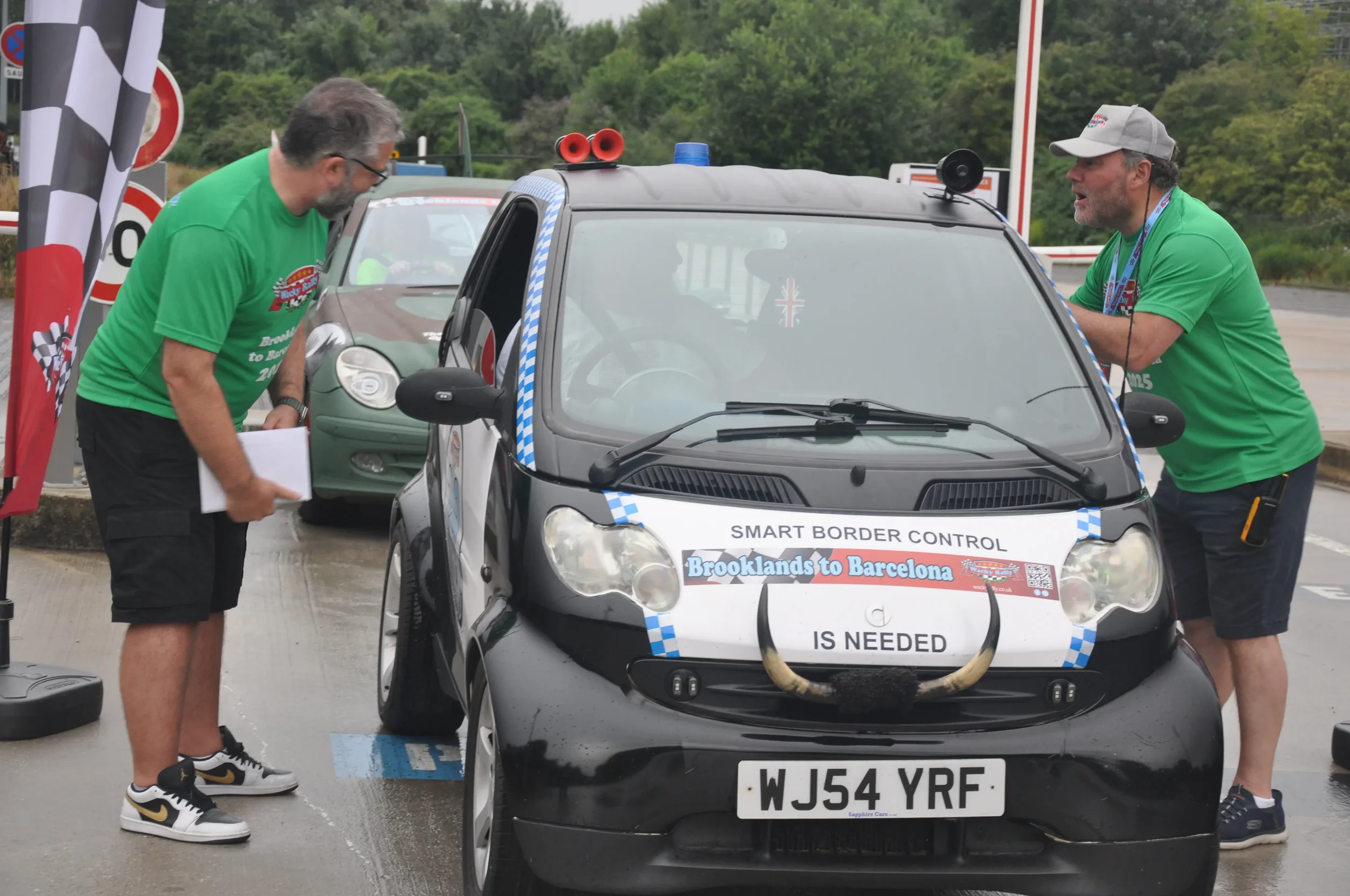 Two men in green shirts at a car border control checkpoint, with a small black car that has a sign on it reading 'SMART BORDER CONTROL' and 'Brooklands to Barcelona IS NEEDED'. The car has a horn on the front, decorated with a bull's horns, and the l