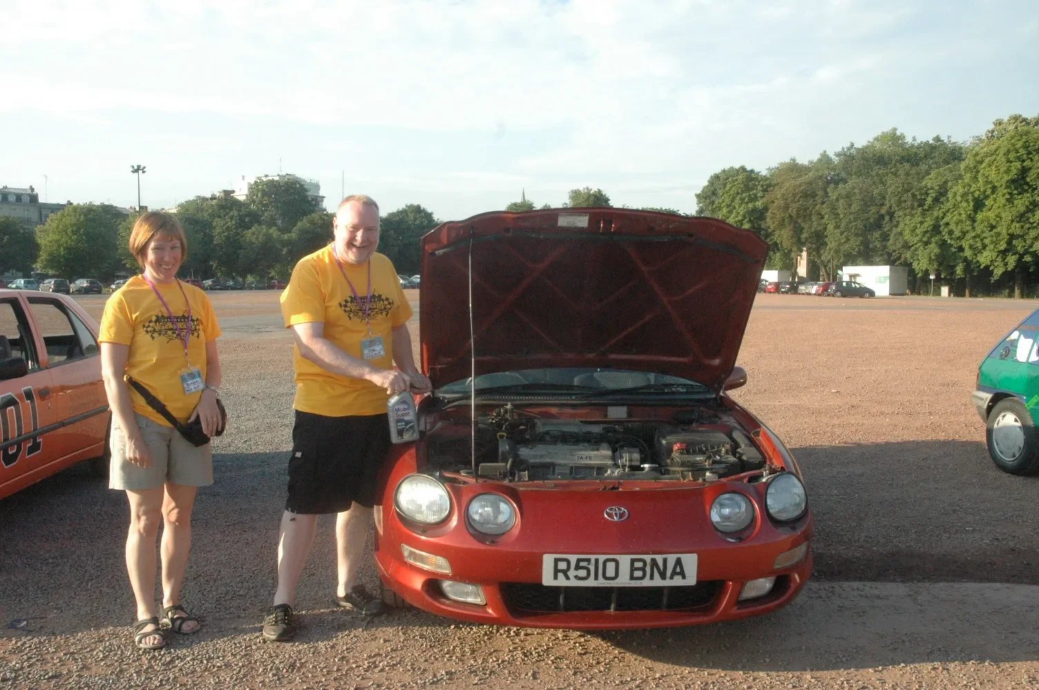 Two smiling people in yellow t-shirts standing next to a red car with its hood open in a parking lot.