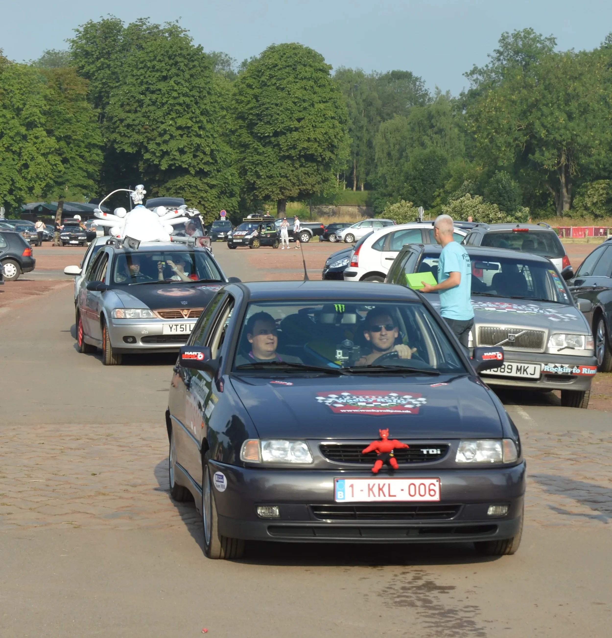 Black compact car with a red plush toy on the front grille, parked in front of other cars at an outdoor event, with two people inside, one wearing sunglasses, and a person standing beside it interacting with the driver.