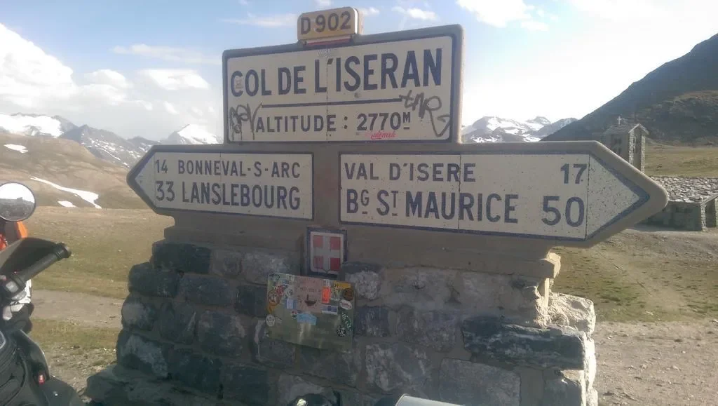 Road sign at Col de l'Iseran indicating directions and distances to nearby locations with snowy mountains in the background.