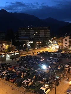 A parking lot filled with cars at dusk, with buildings and mountains in the background under a cloudy sky.