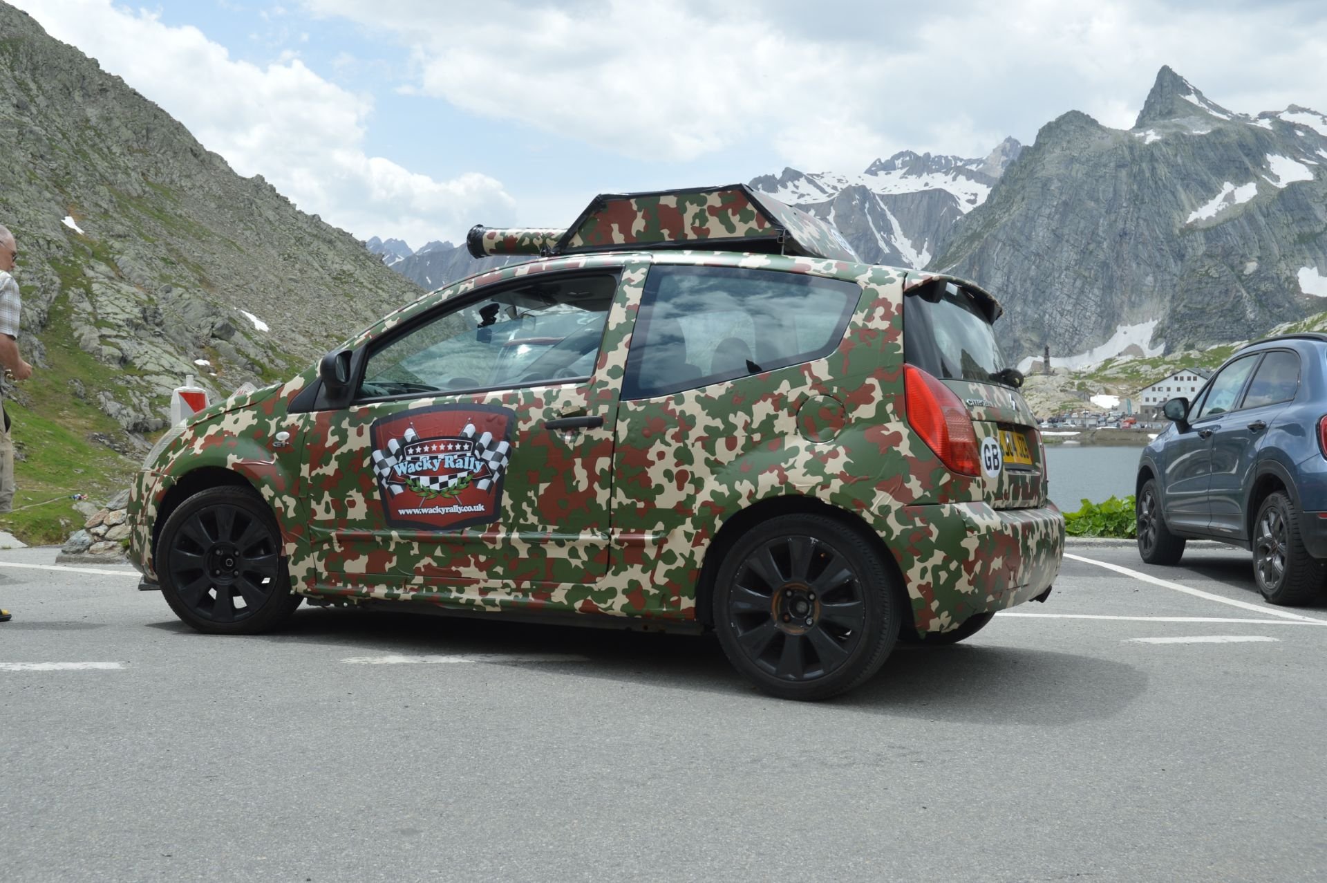 Camouflaged car with rally stickers parked near a mountain lake with snowy peaks in the background.