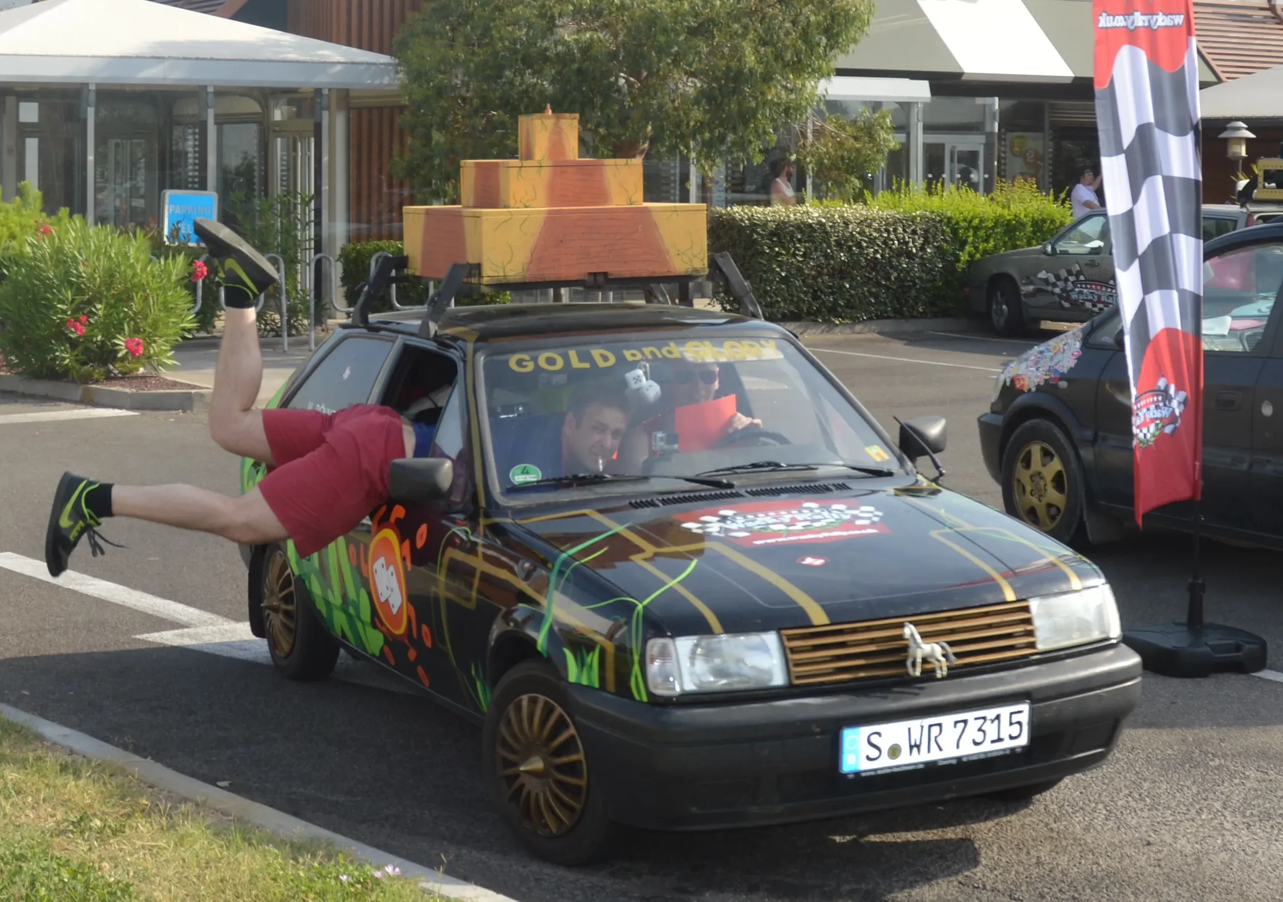 A decorated black car with racing-themed graphics and a large wooden crate on top, parked in a lot. Two people are in the front seats, one leaning outside the car with legs in the air, smiling.