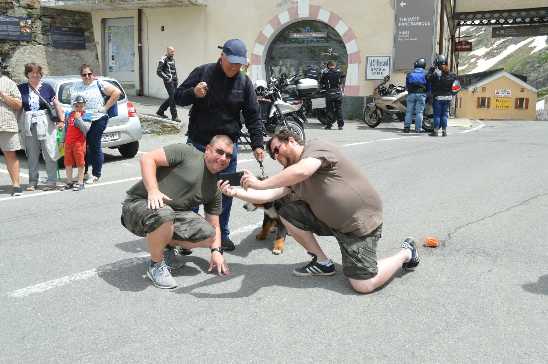 Two men kneeling and taking a selfie with a dog on a leash, while a woman stands nearby and others watch and take pictures in a mountain roadside area.