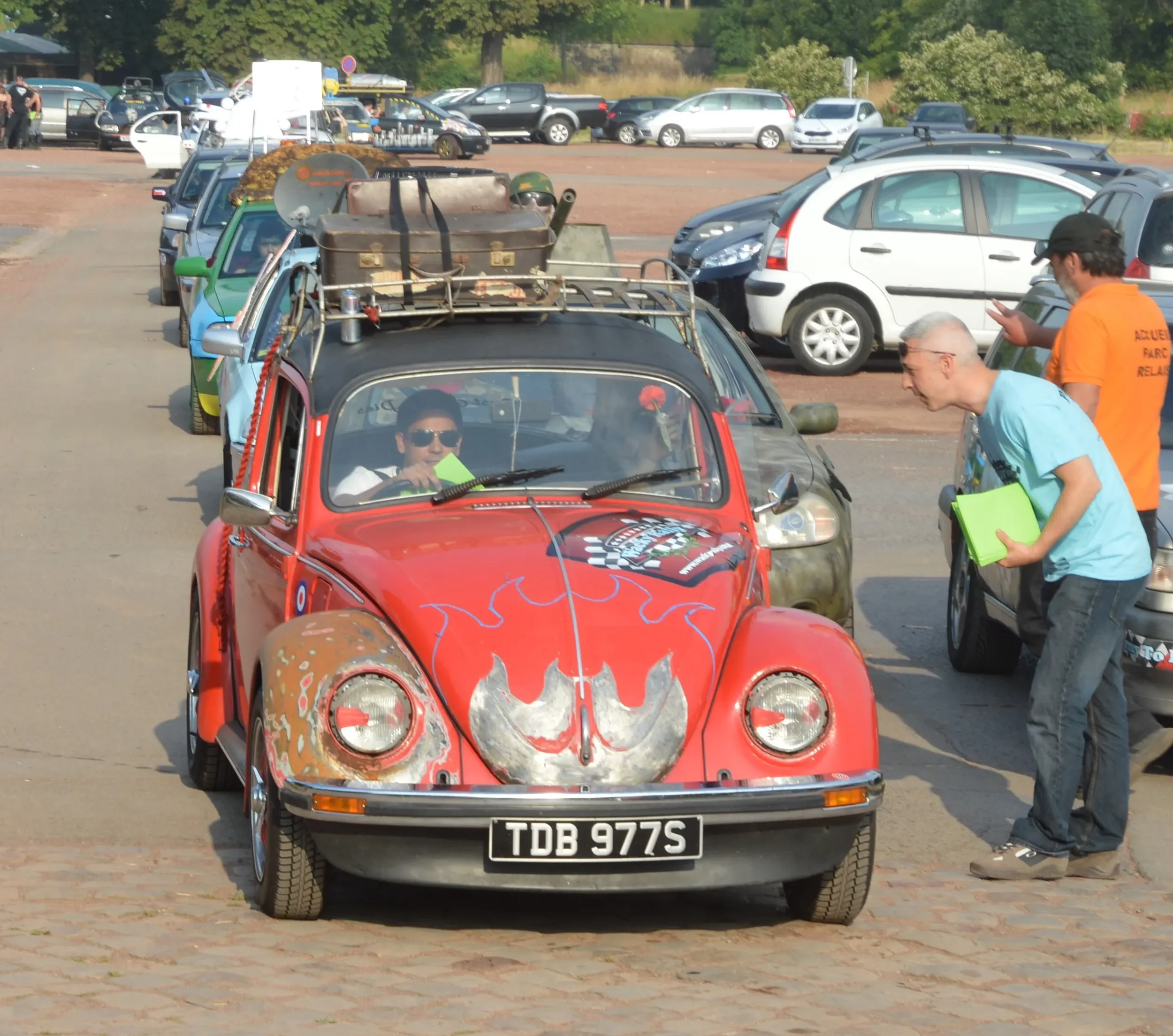 A red vintage Volkswagen Beetle decorated with Batman-themed artwork on the hood, with three men interacting nearby in a crowded parking lot under sunny weather.
