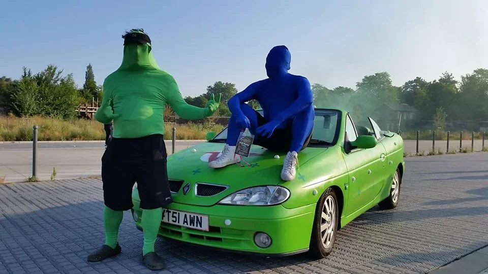 Two people wearing full-body colored suits, one in green and one in blue. The green-suited person is standing and talking to the seated blue-suited person on a bright green convertible car. The blue-suited person is sitting on the car's hood, wearing
