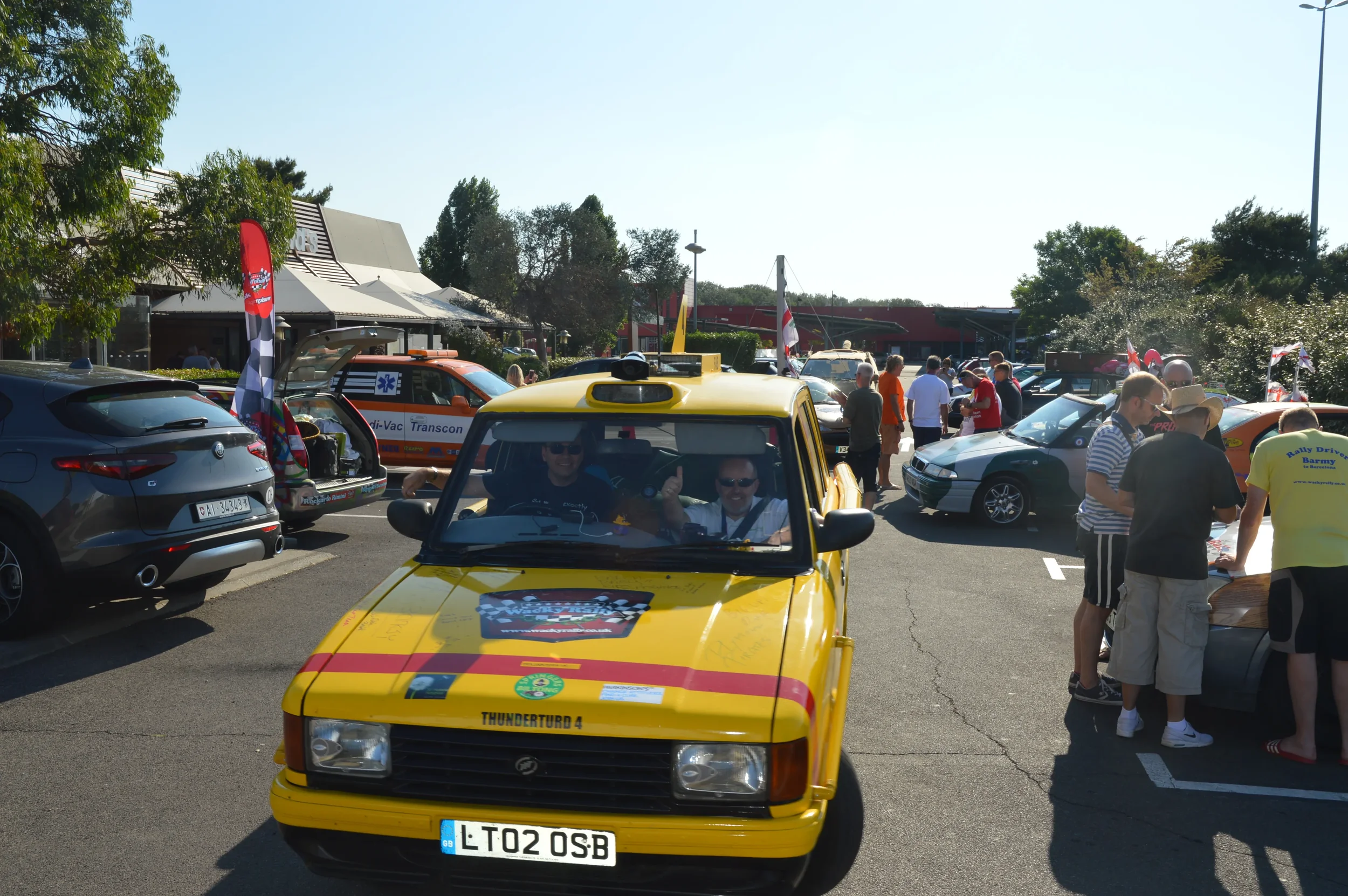 A yellow vintage car with two smiling men inside, giving a thumbs up, parked in a busy parking lot during a car event with people and various cars around.