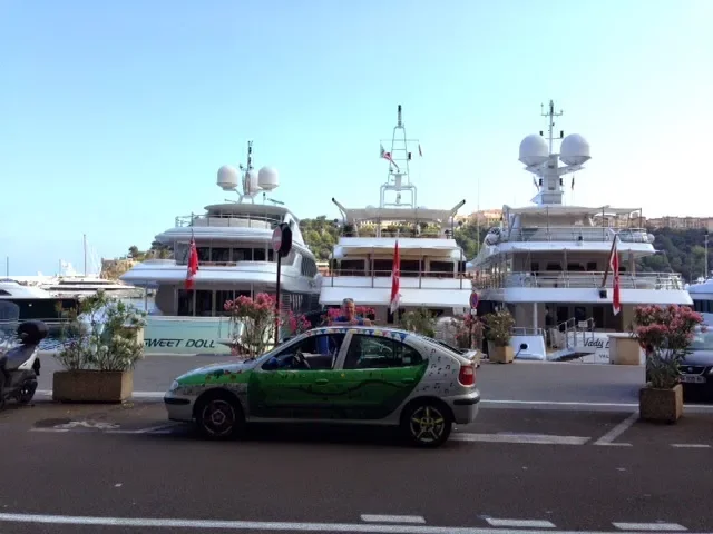 Three luxury yachts docked at a marina with a green and silver car parked in front