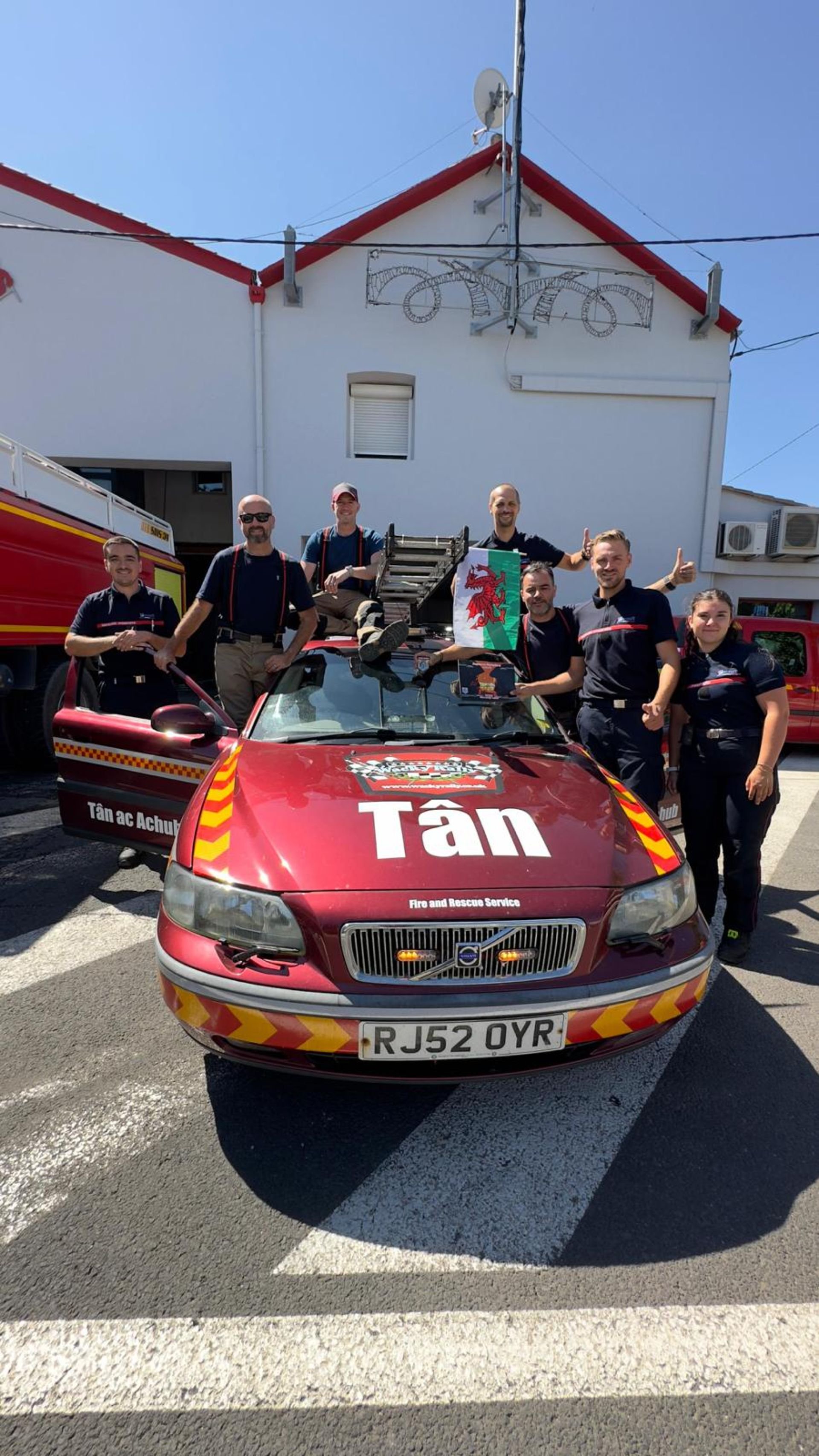 Group of firefighters standing around a red fire rescue car, some giving thumbs up, in front of a white building on a sunny day.