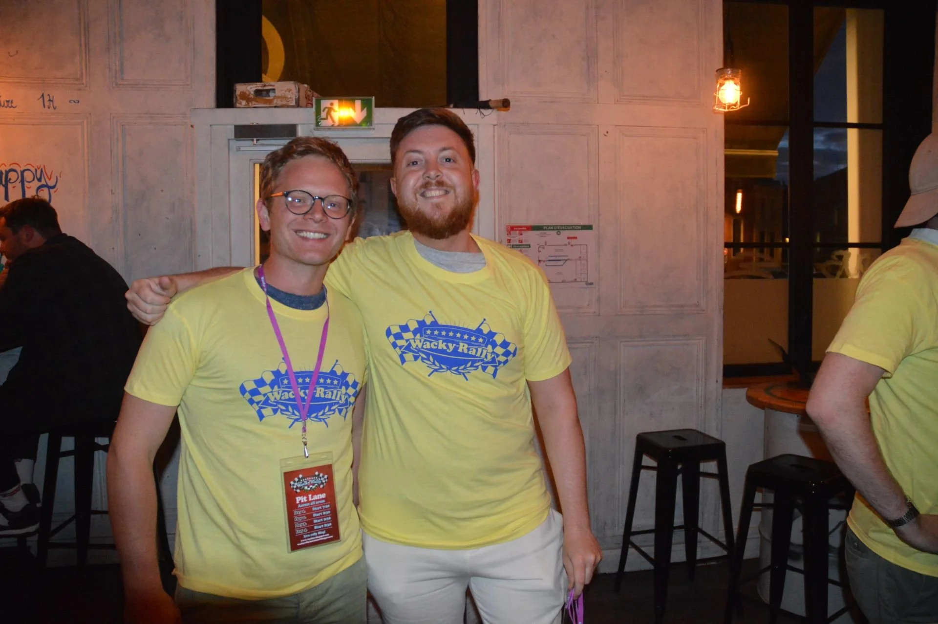 Two men wearing yellow t-shirts with 'Wacky Rally' logos, smiling and posing with their arms around each other in a dimly lit indoor setting.
