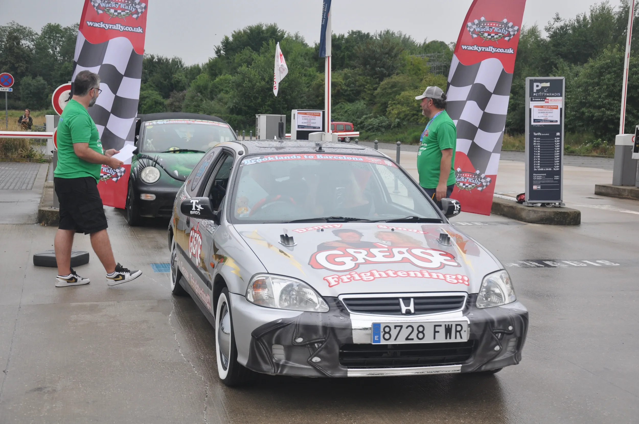 A silver Honda car with racing graphics and the words 'GREAT! Fighting!!' on the hood, parked at a rally event with two men in green shirts standing nearby. There is a second car behind it, and banners with checkered flags and the website 'wackyrally