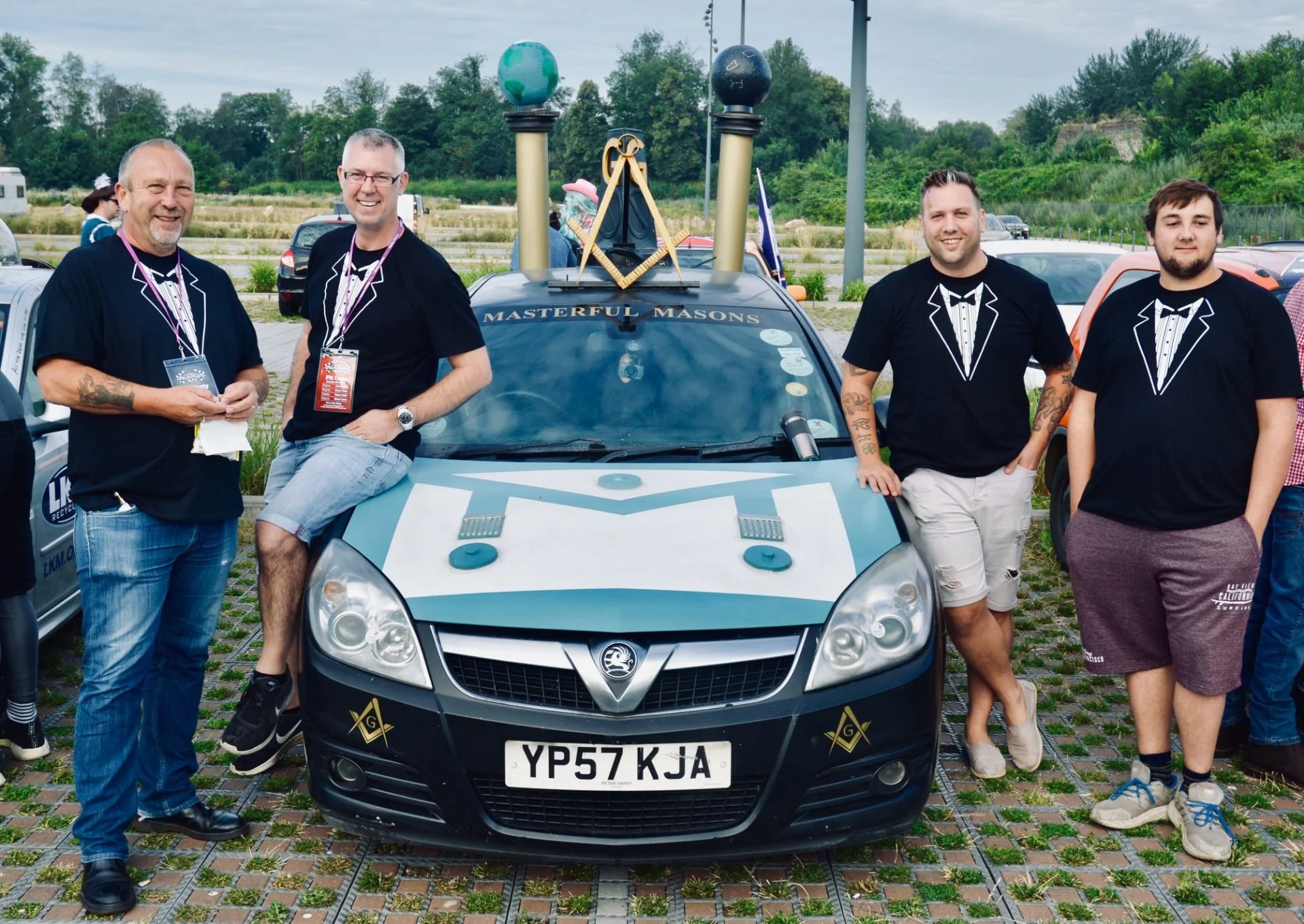 Group of four men standing around a decorated police car with Masonic symbols, at an outdoor event, wearing black shirts with tuxedo prints, some with tattoos and casual shorts.