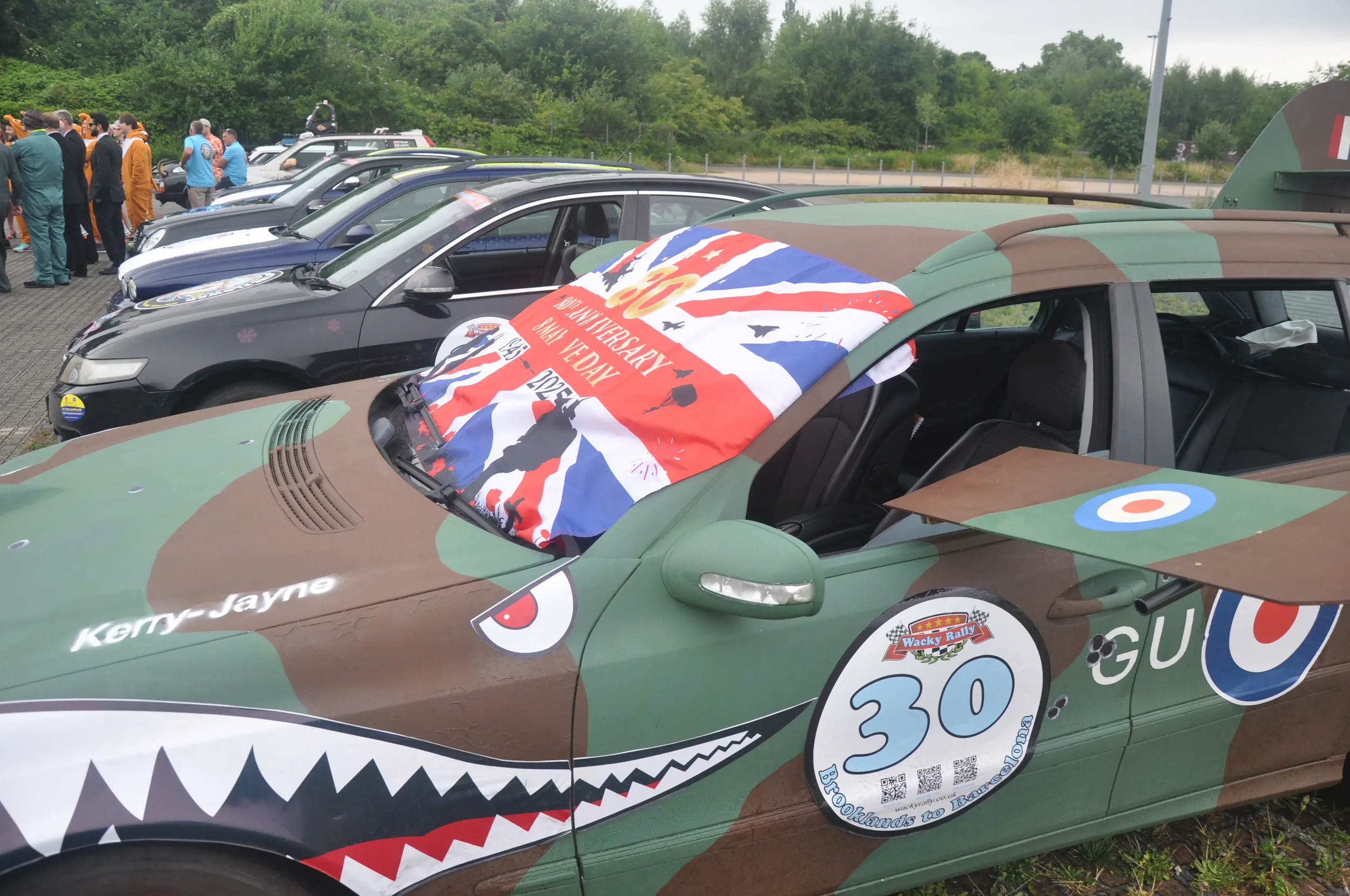 A line of cars parked outdoors, featuring a camouflage-patterned rally car with a large shark mouth graphic, a number 30, and a patriotic flag-style banner draped over the windshield. People are gathered in the background.