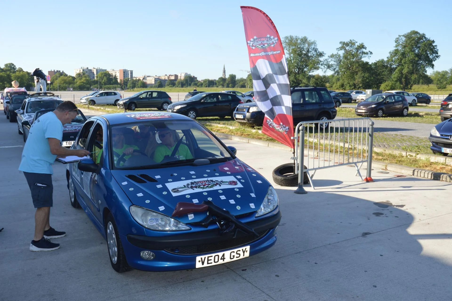 Blue car with a deformation on the hood and the number plate VE04 GSY, parked at a racing event. A person in a white shirt is writing on a clipboard near the car. Two people in bright green shirts are sitting inside the car. A red racing flag, a barr