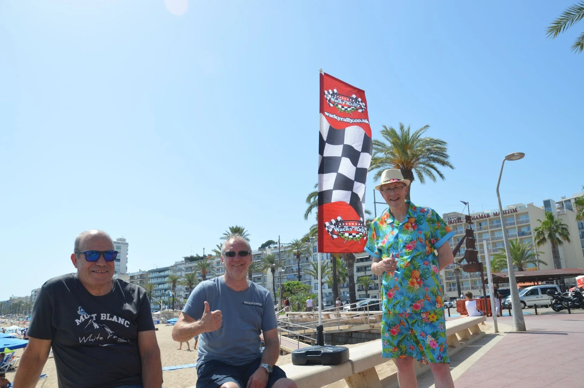 Three men sitting and standing near a beach promenade with palm trees, buildings, a hotel, and a racing flag in the background. Two of them are sitting on a concrete ledge, one giving a thumbs up, and the third standing in a colorful Hawaiian shirt, 