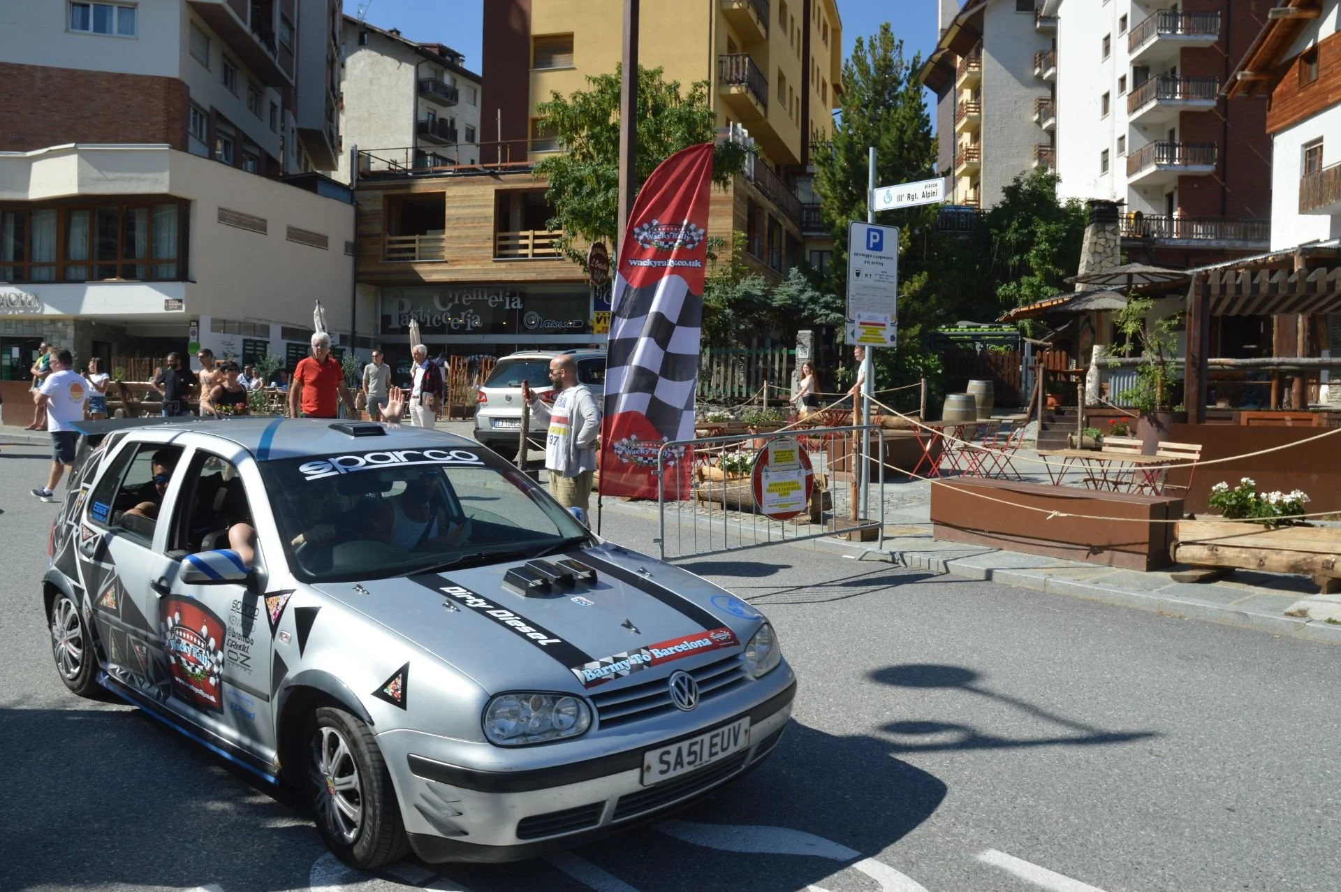 A modified Volkswagen car with rally decals and sponsors parked on a city street during a racing event, with people walking and some onlookers in the background.