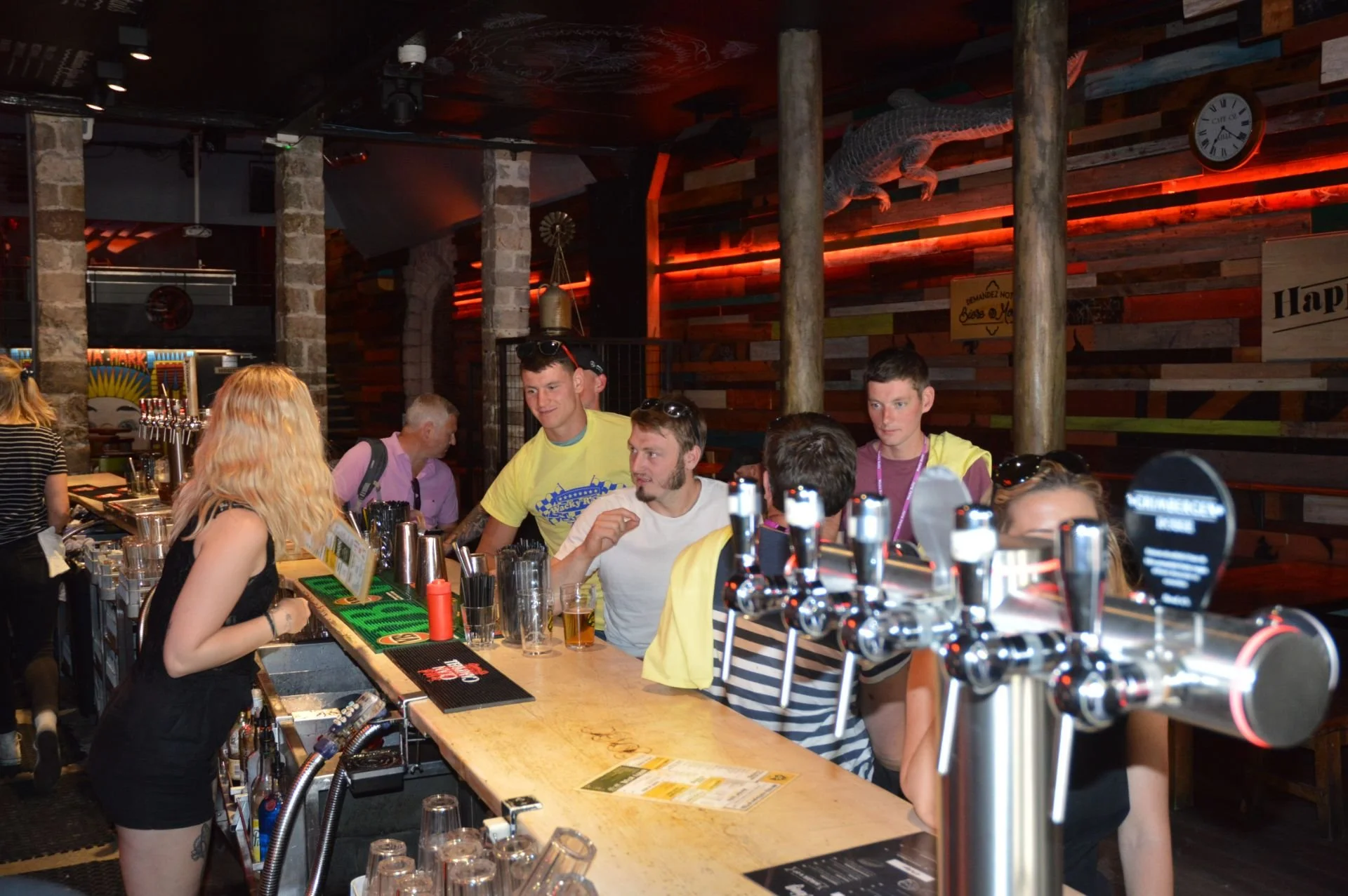 People gathered at a bar counter with glasses, beer taps, and bar menus inside a dimly lit setting with colorful decor, including a crocodile sculpture on the wall.
